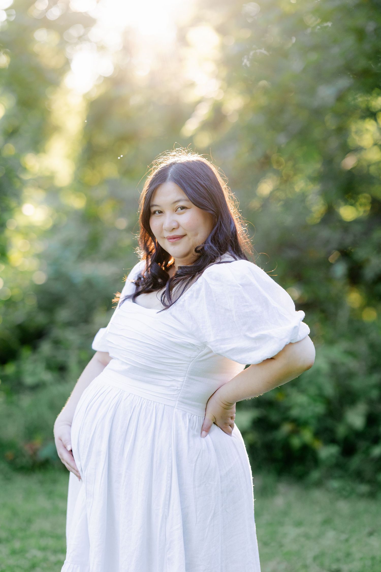 Woman in a flowing white dress poses in a sunlit garden setting.