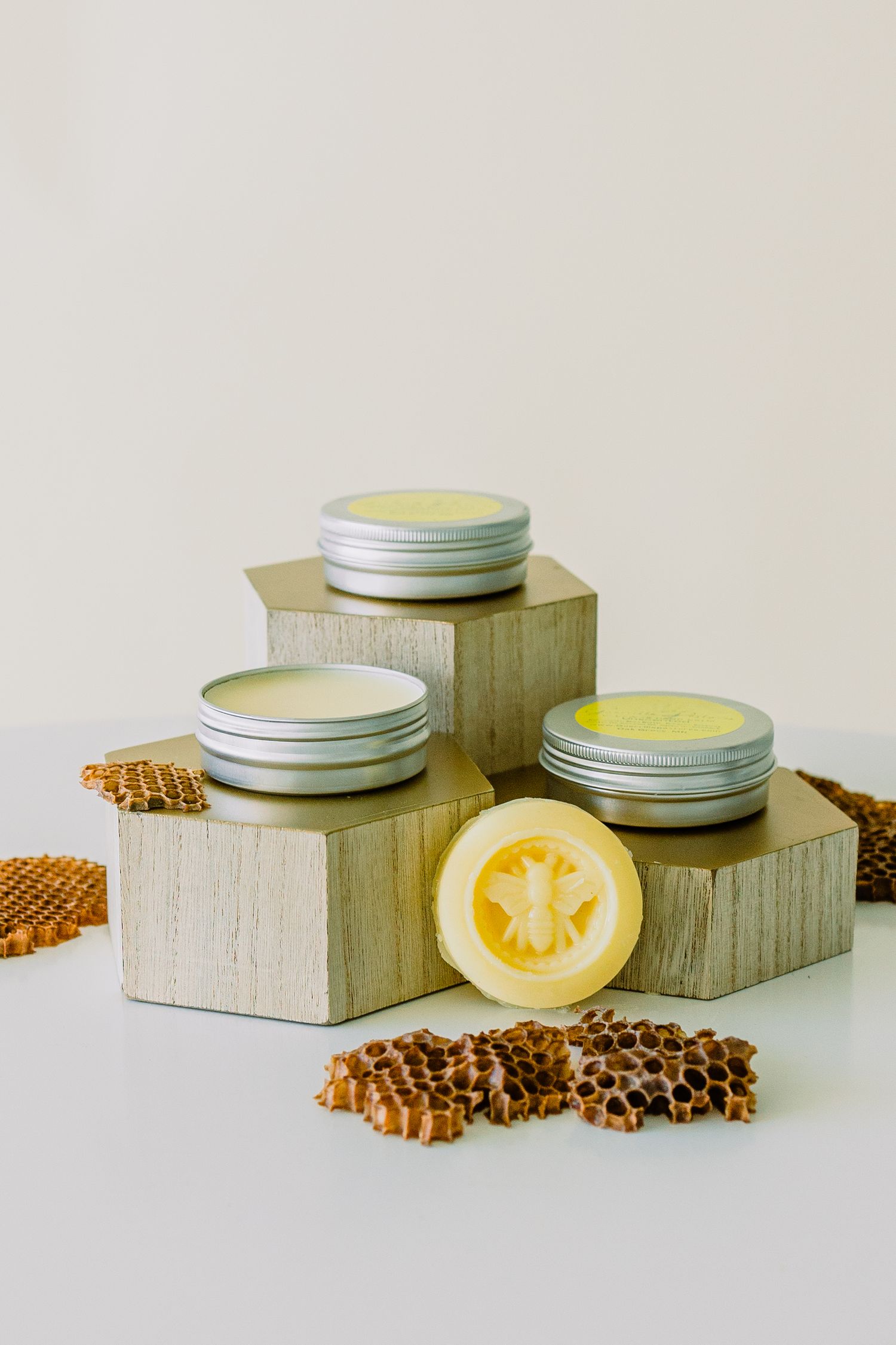 Three metal tins of homemade beauty products displayed on wooden blocks with honeycomb pieces and citrus slice decorations.