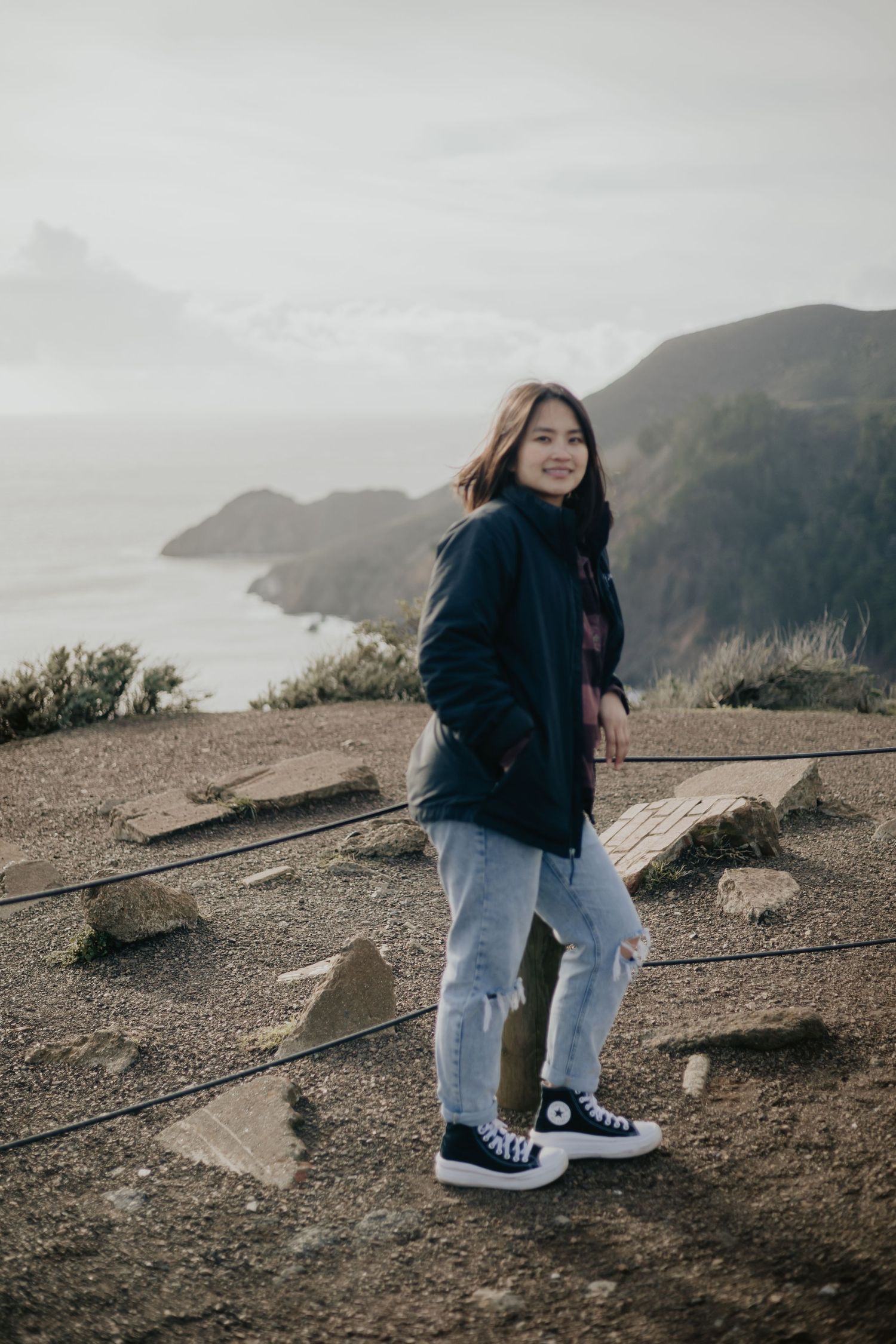 Person in a navy jacket and jeans stands on a cliff overlooking a misty coastal view with dramatic mountains.