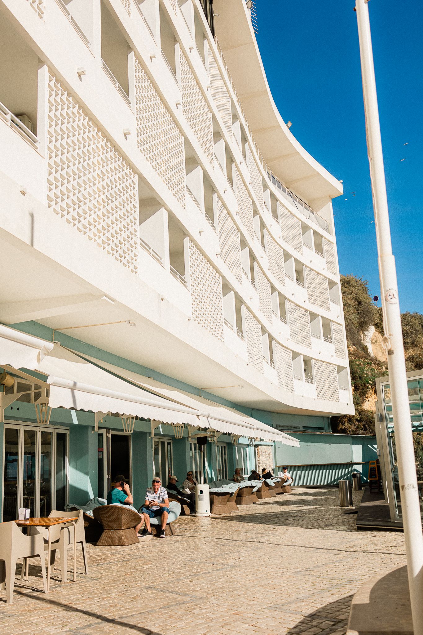 Modern white hotel building with latticed balconies and outdoor seating area under bright blue sky.