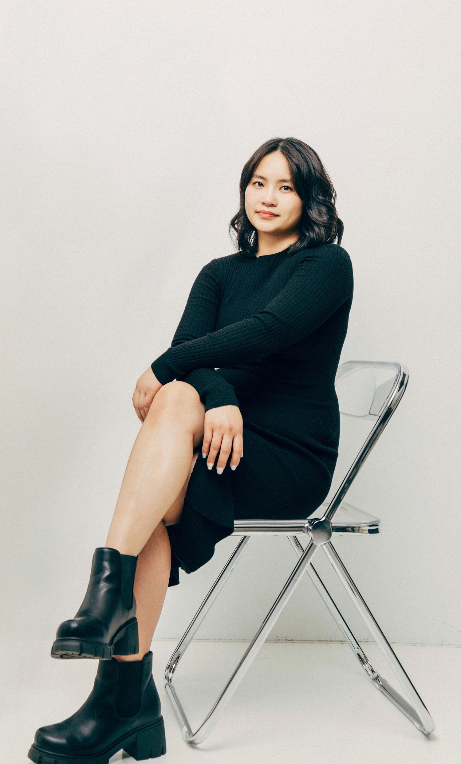 Someone wearing a black sweater dress and chunky boots sits gracefully on a modern folding chair against a plain white background.