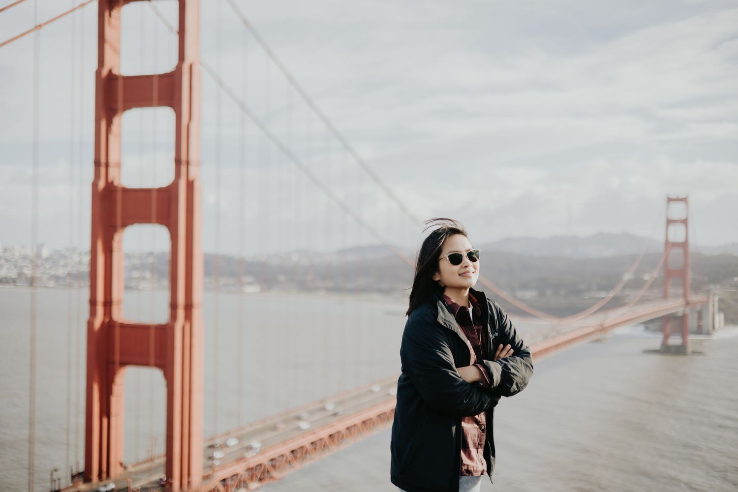 Tourist in black jacket stands with arms crossed near the iconic Golden Gate Bridge in San Francisco on a cloudy day.