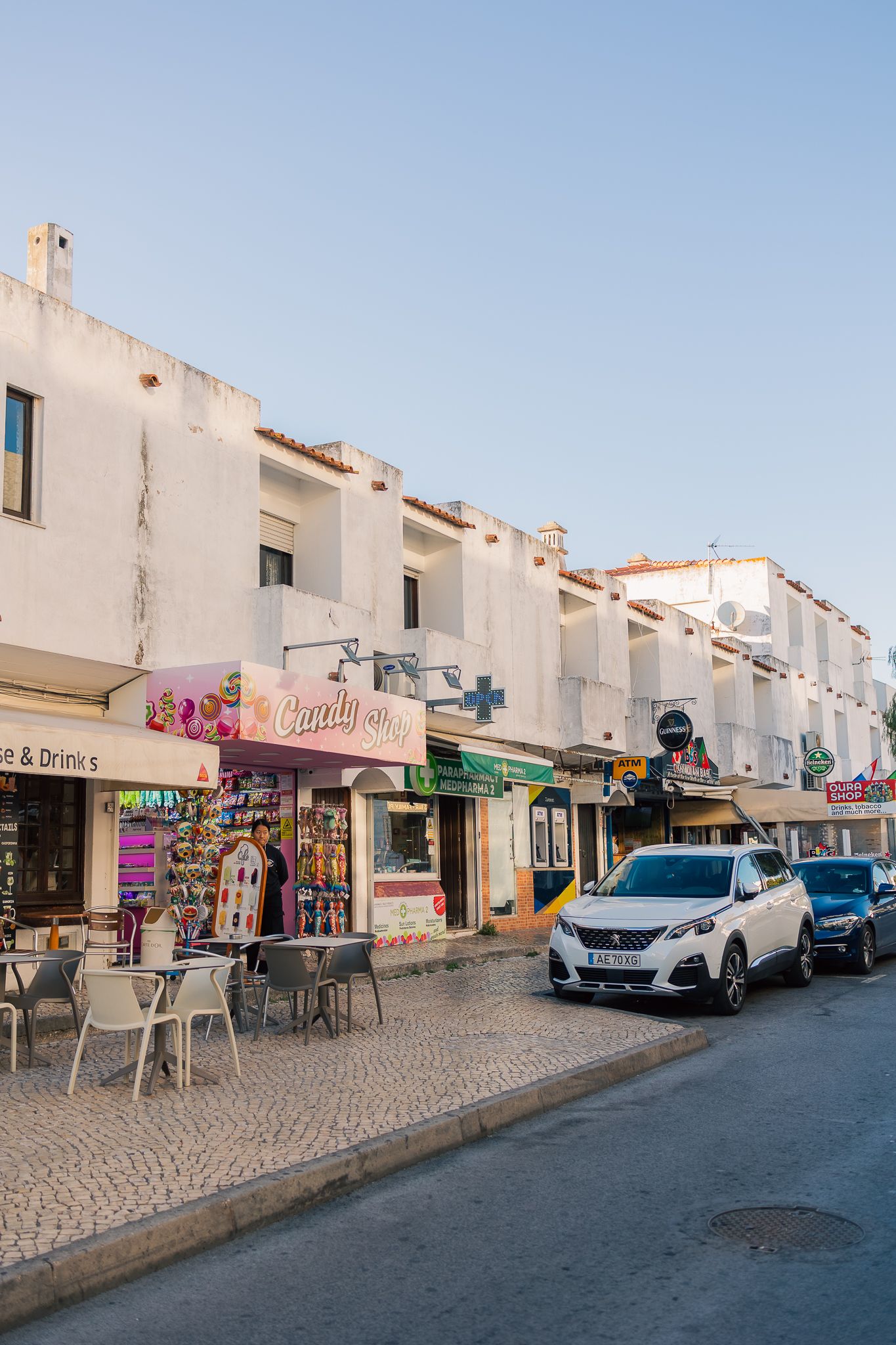A charming street in Portugal with white buildings, shops, and parked cars along a cobblestone road.