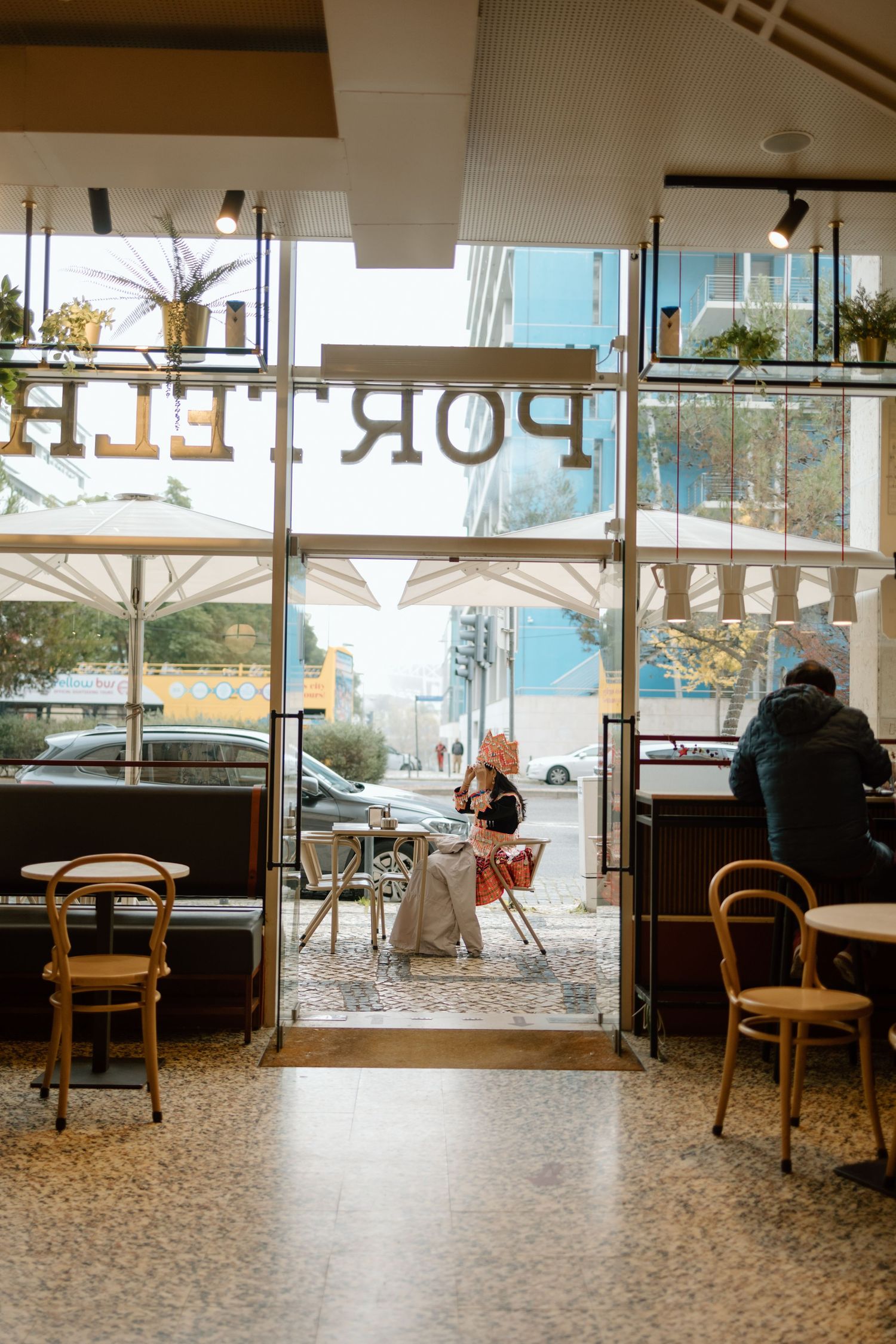 View through cafe window showing wooden chairs and terrazzo floor with downtown buildings visible in background.