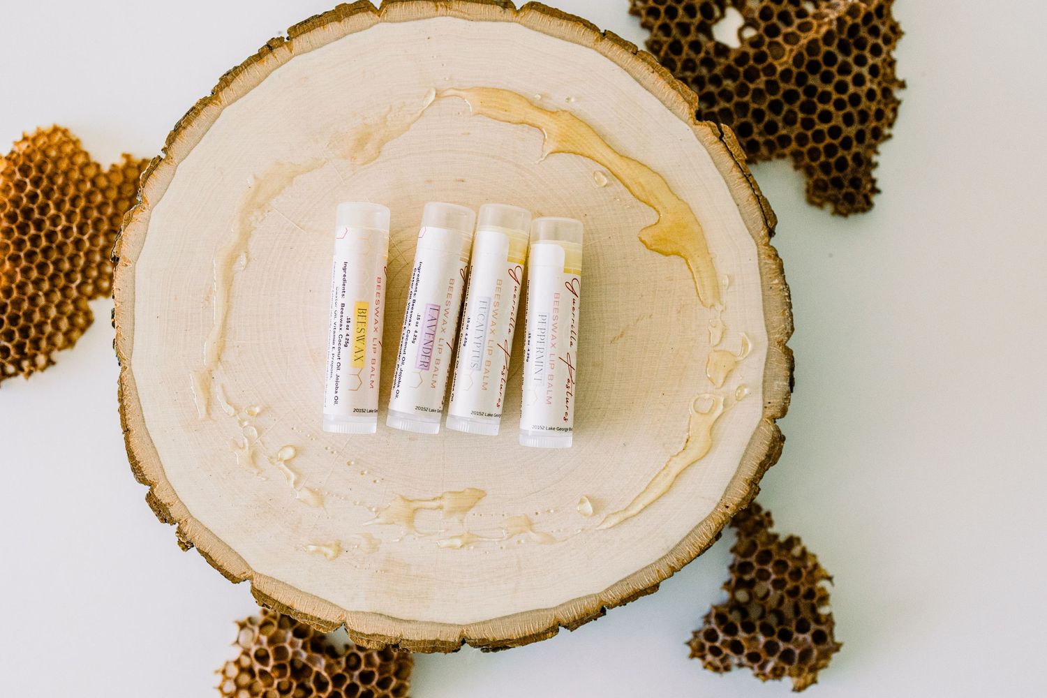 Small white lip balm tubes arranged on a round wooden slice with decorative dried peppercorns scattered around.