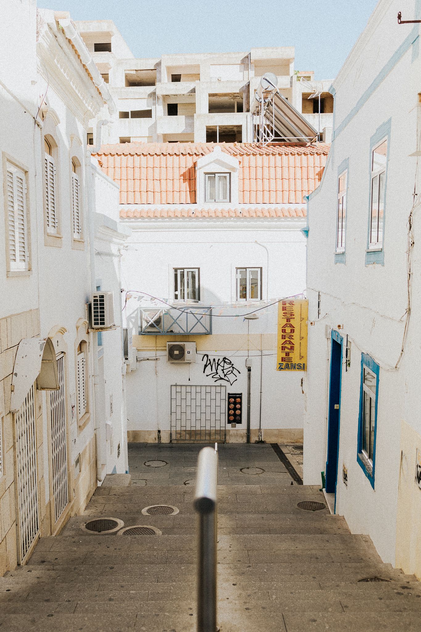 A narrow alleyway in a Mediterranean town with white buildings, terra cotta roofs, and blue doors.