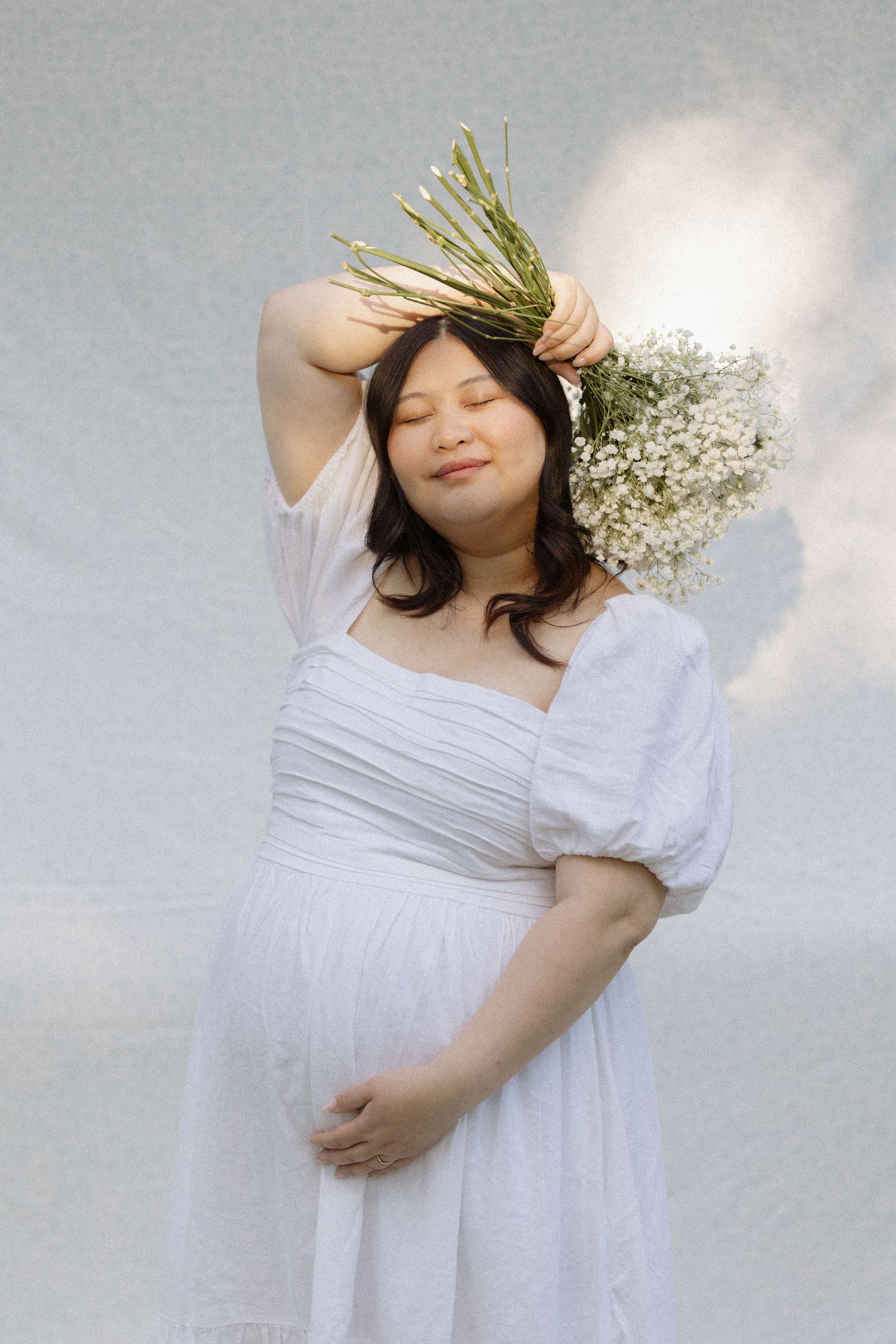 A person in a flowing white dress poses against a light background with baby's breath flowers and pine branches in their hair.