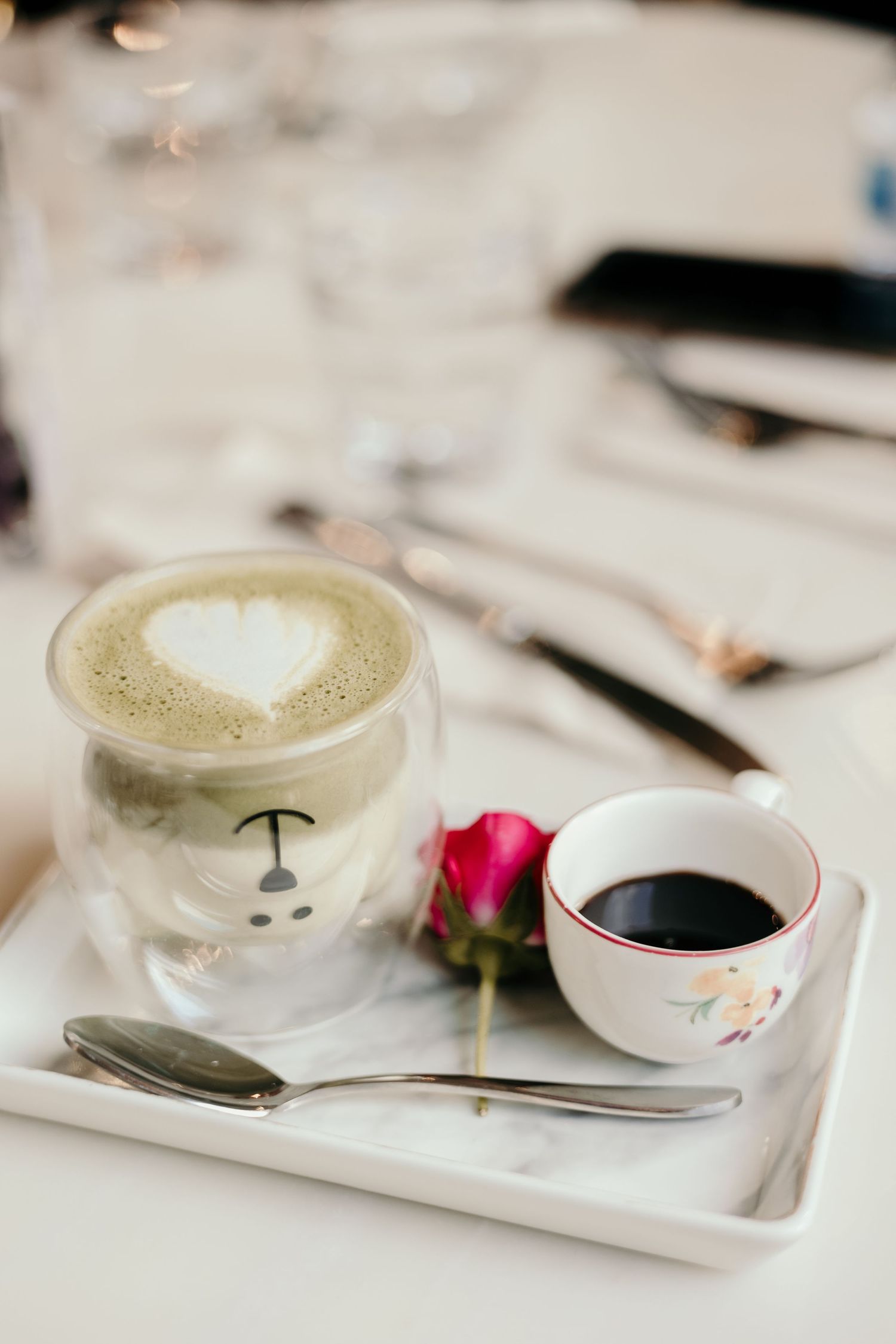 Two coffee cups on a white tray with a red rose, one featuring latte art heart design.