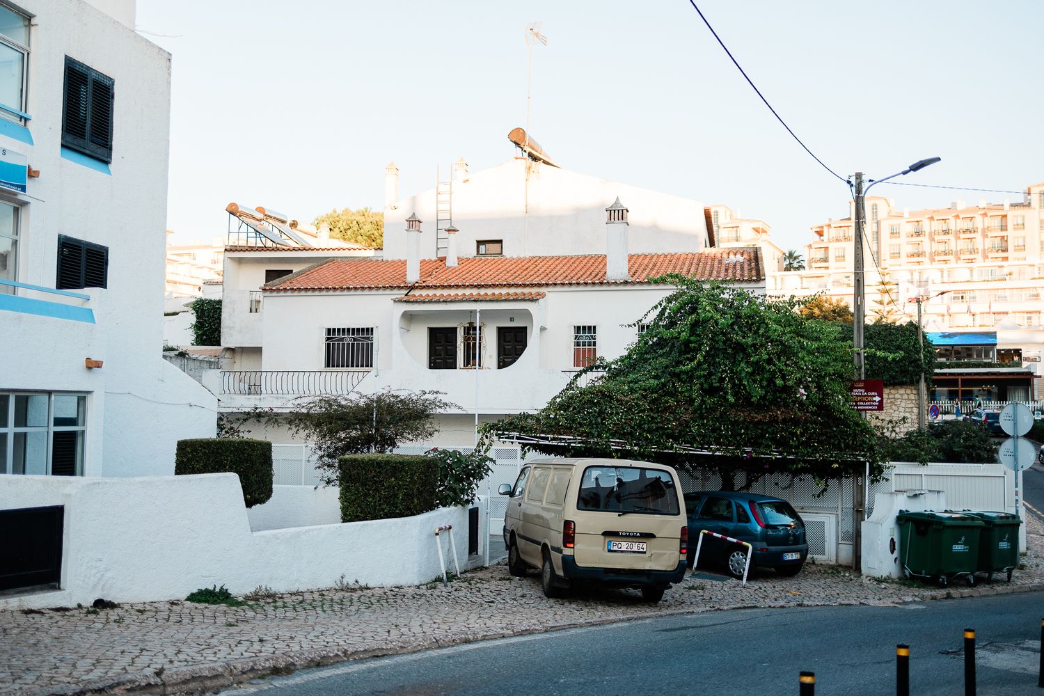 A white van and blue car parked outside whitewashed buildings in a coastal Mediterranean town.