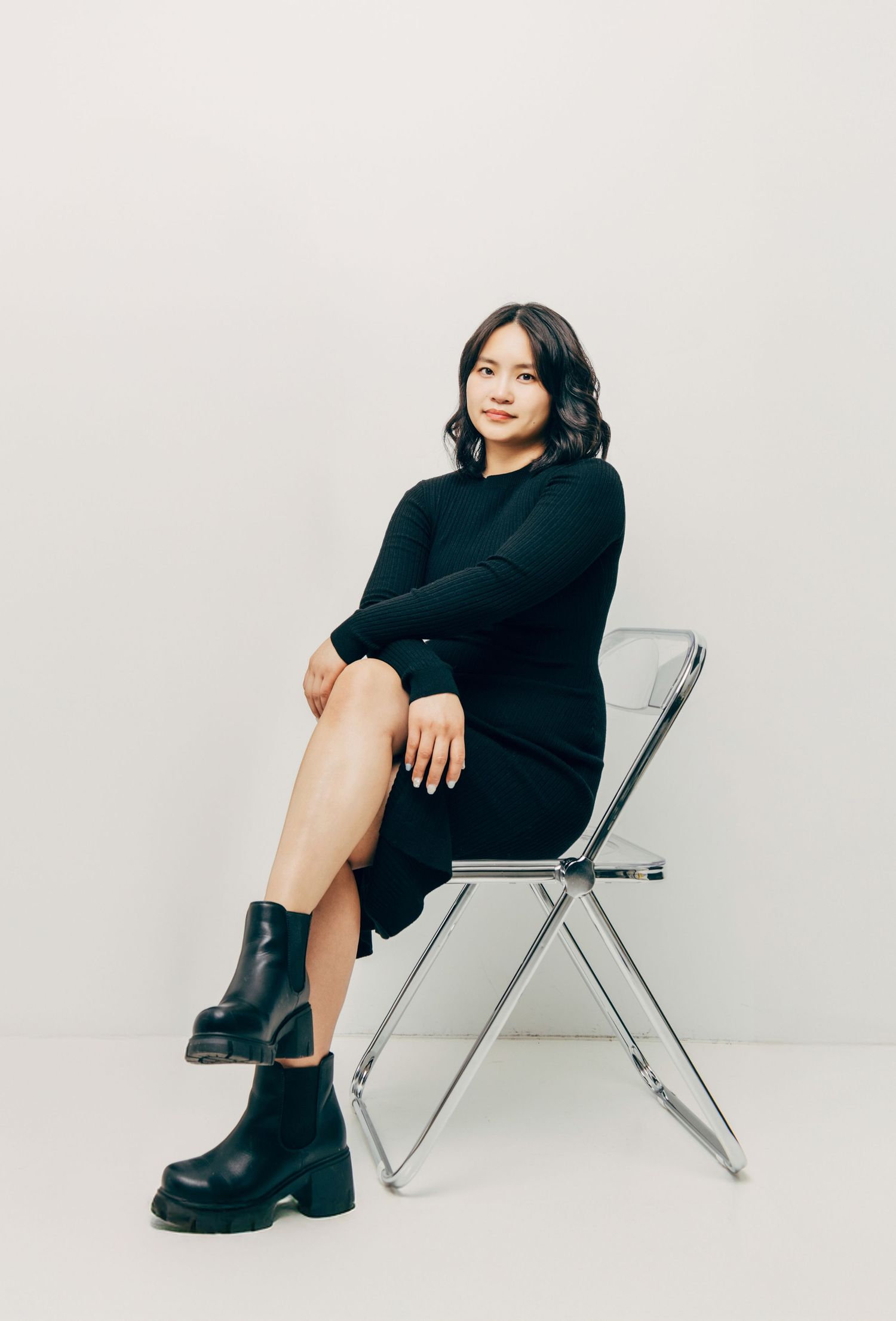 Someone in a black knit dress sits elegantly on a metal folding chair wearing chunky black boots against a light background.