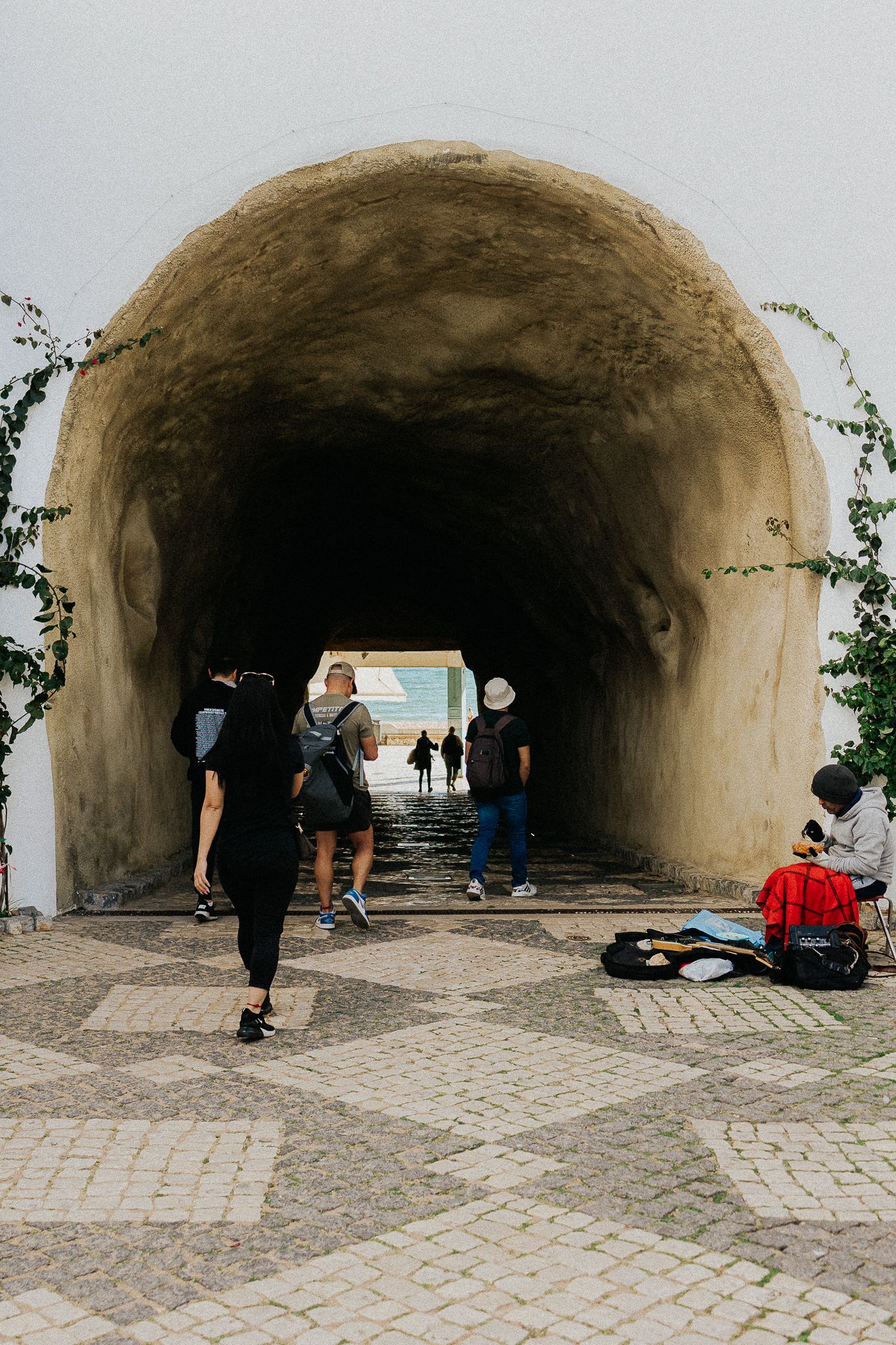Pedestrians walk through a curved stone tunnel entrance with cobblestone pavement and a street musician performing.