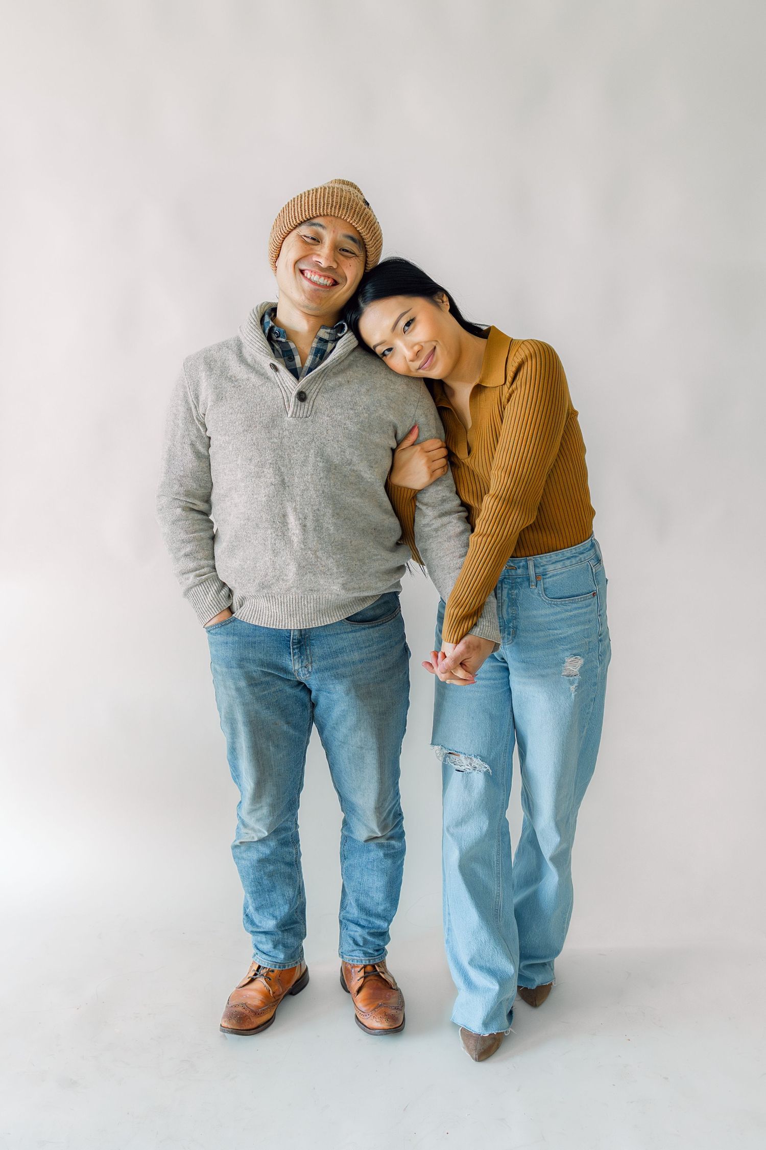 Couple in casual sweaters and jeans standing together against white background in intimate embrace.