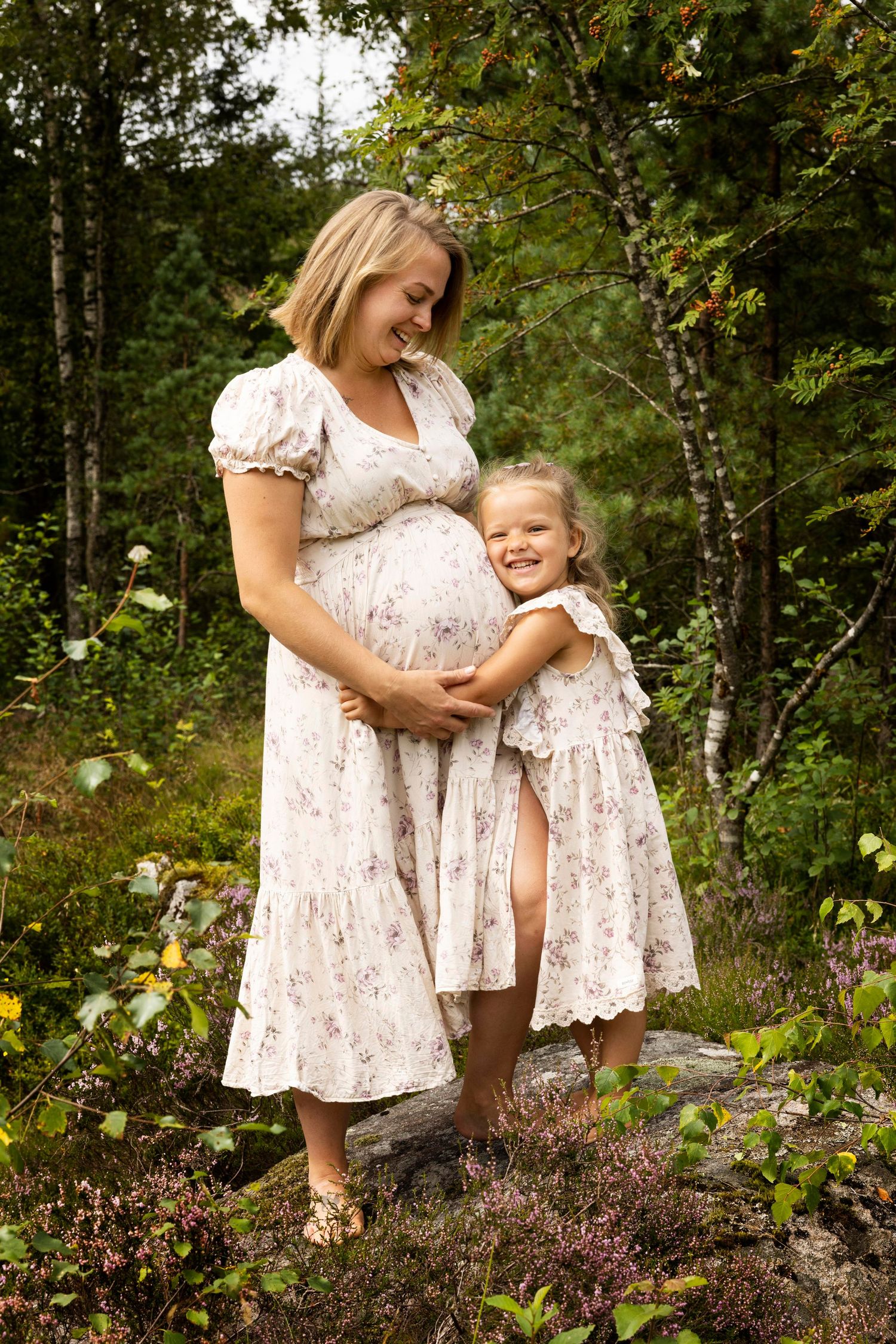 Gravidmamma og datter i like sommerkjoler holder rundt hverandre og ler, i skogen i Lyngdal en varm sommerdag