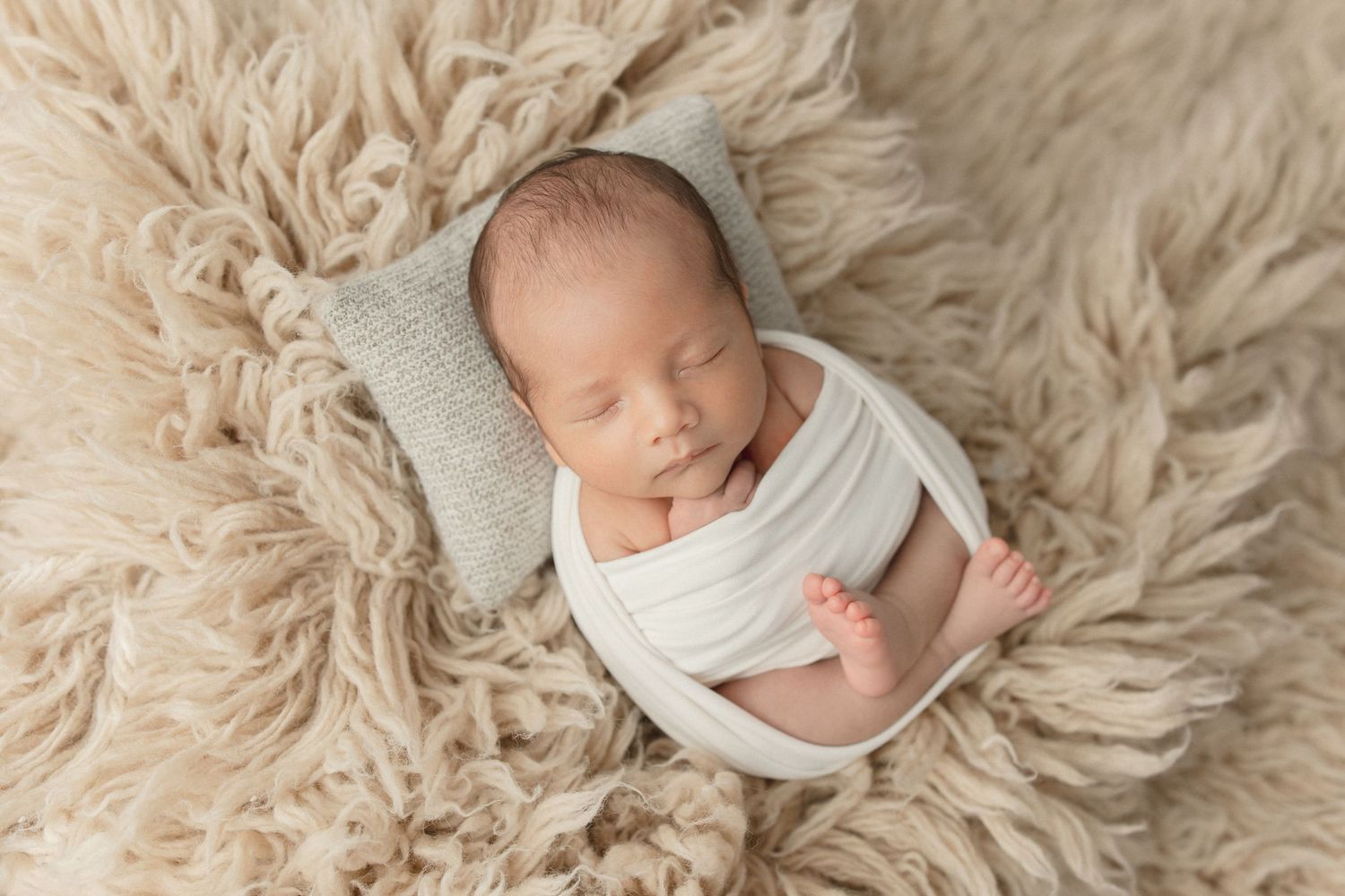 Sleeping newborn wrapped in white swaddle lying on fluffy beige fur backdrop during professional photo session.