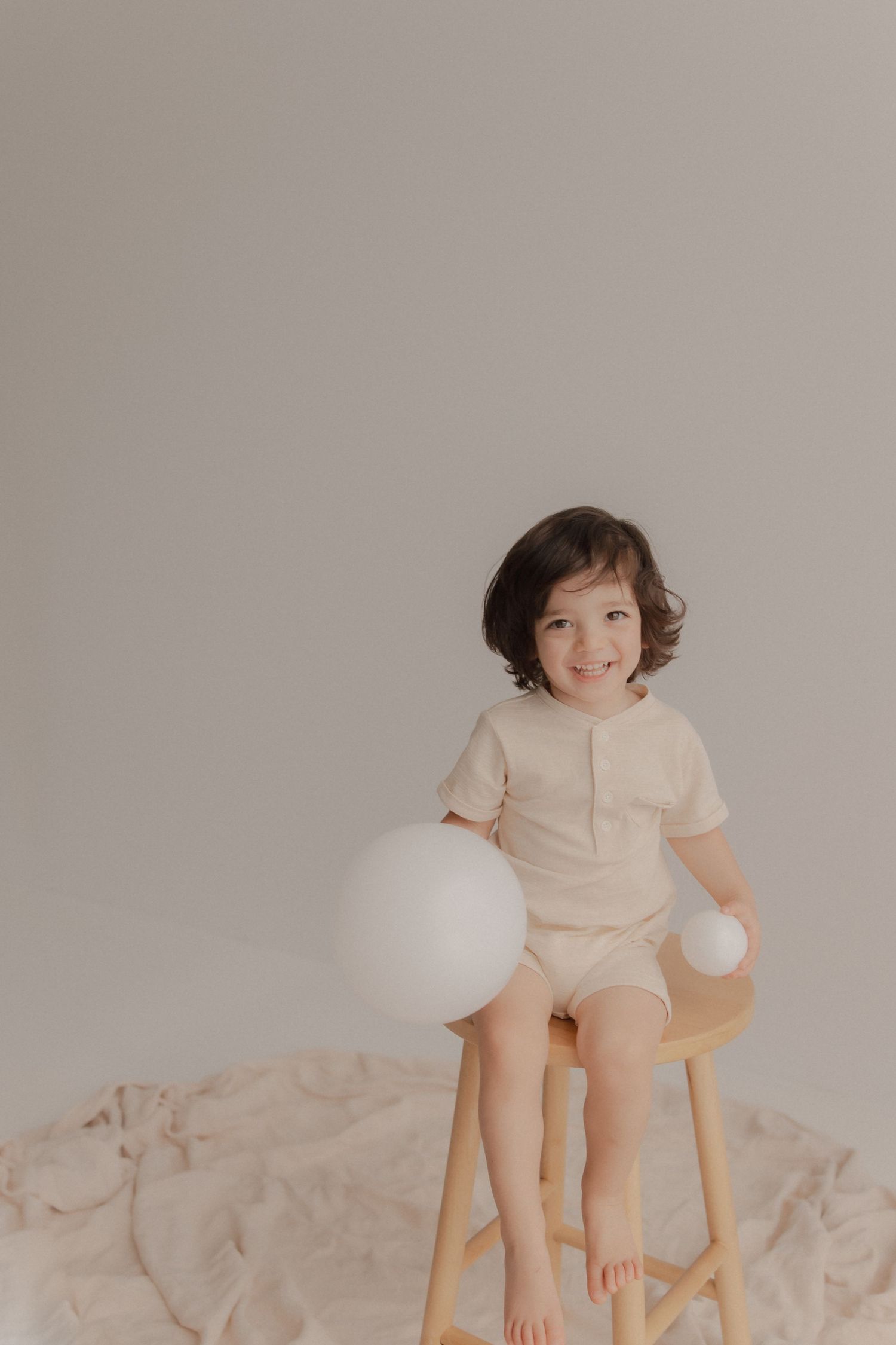 Child in beige outfit sits on wooden stool with white balloon against neutral backdrop for minimalist portrait.