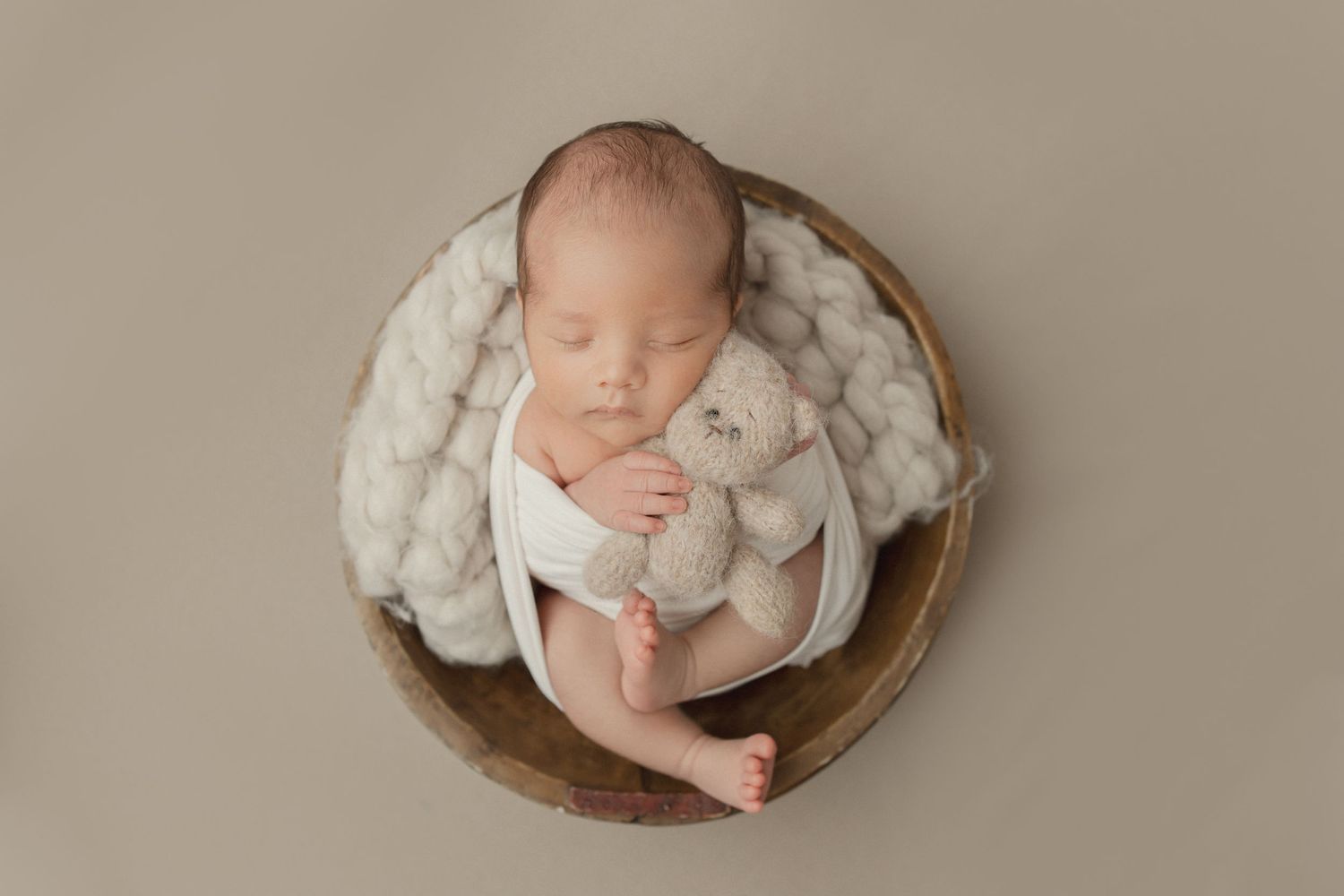 Sleeping newborn bundled in white wrap lies curled in round wooden bowl on neutral beige backdrop.