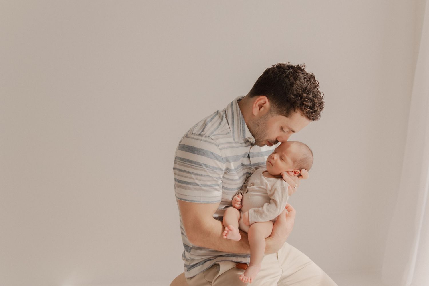 Gentle parental moment captured in soft natural light against neutral background showing tender embrace with baby.