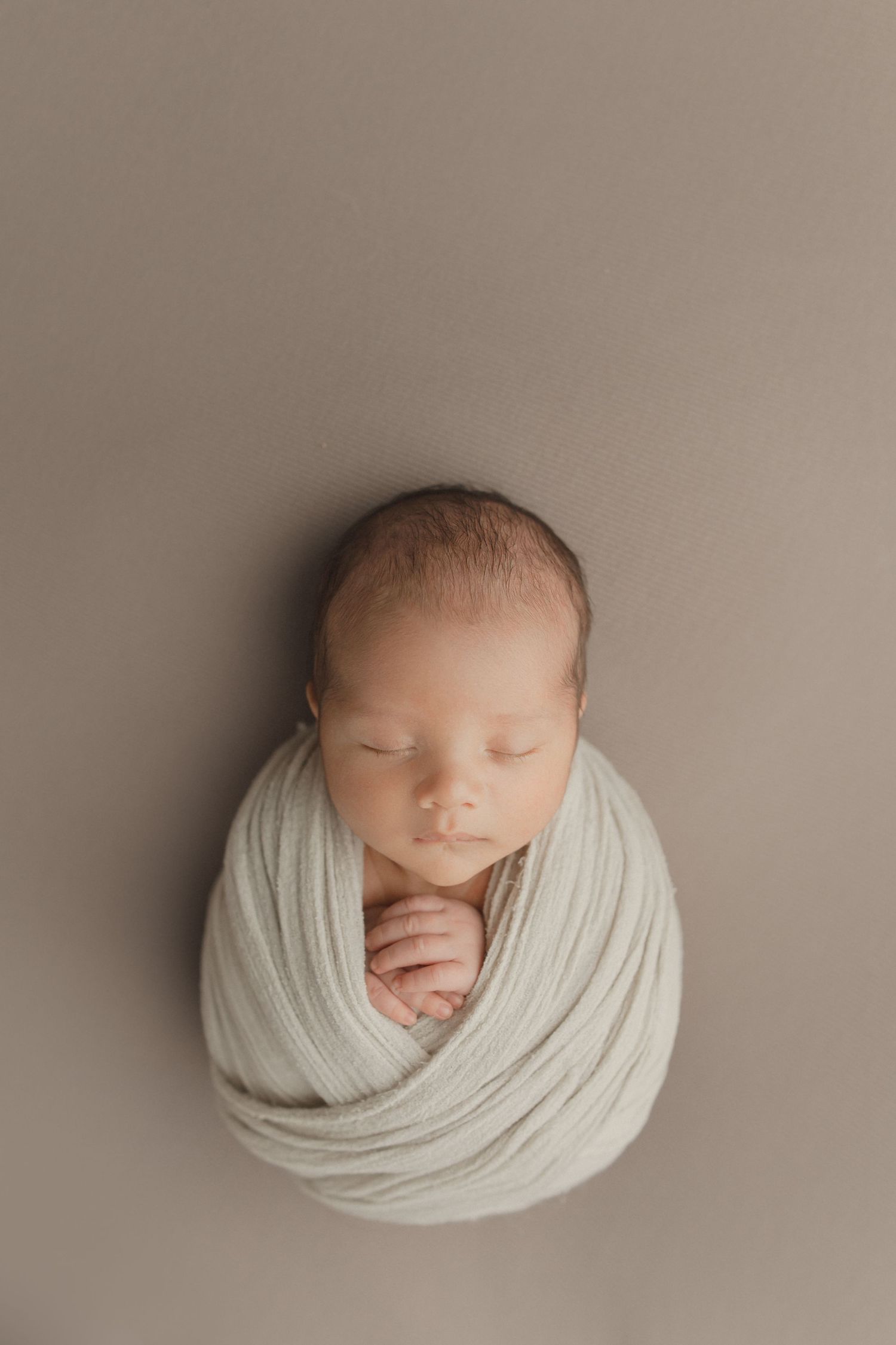 Newborn baby peacefully sleeping while wrapped in soft white fabric against neutral beige background.