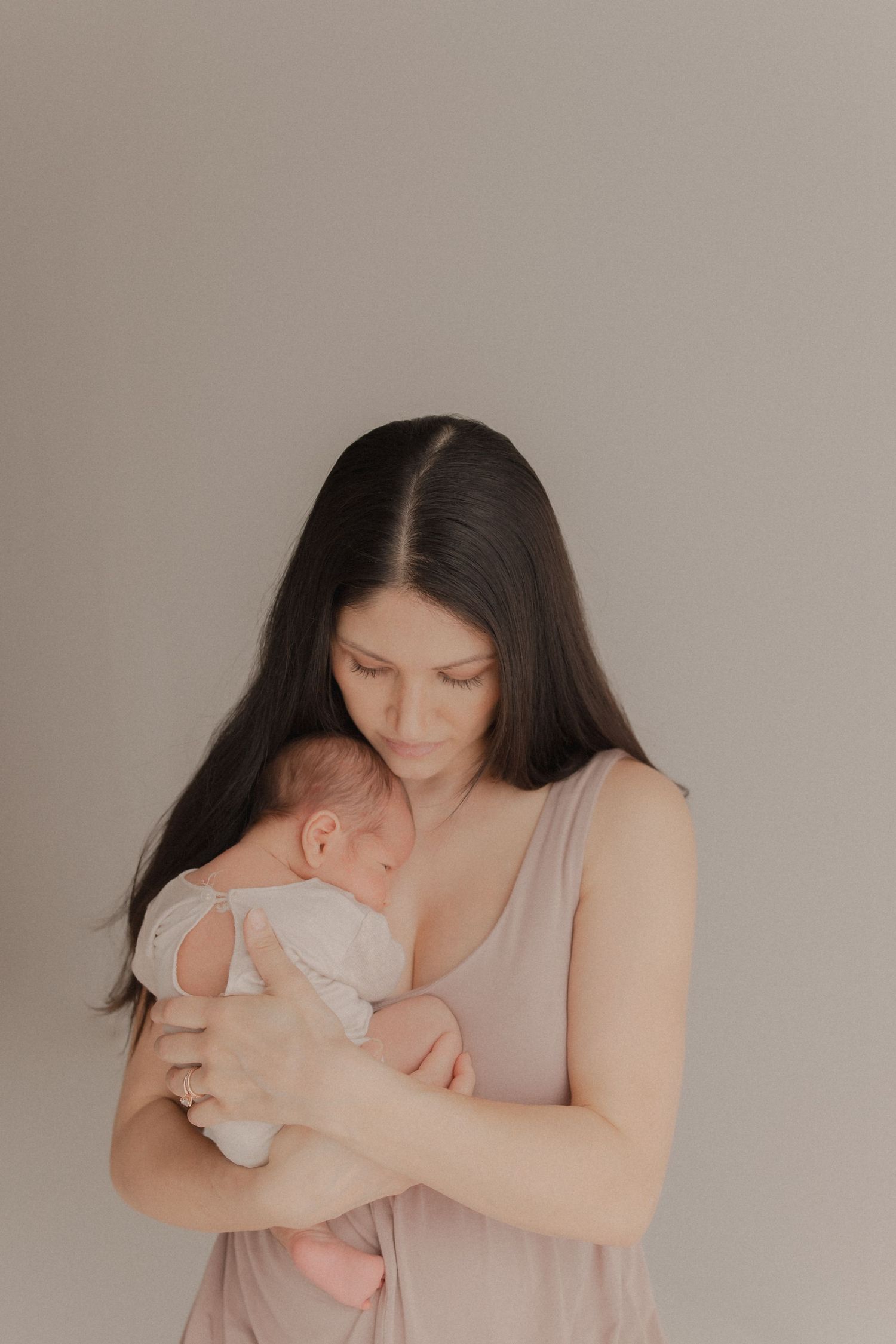 Mother in pink dress tenderly holding sleeping newborn against neutral beige background.