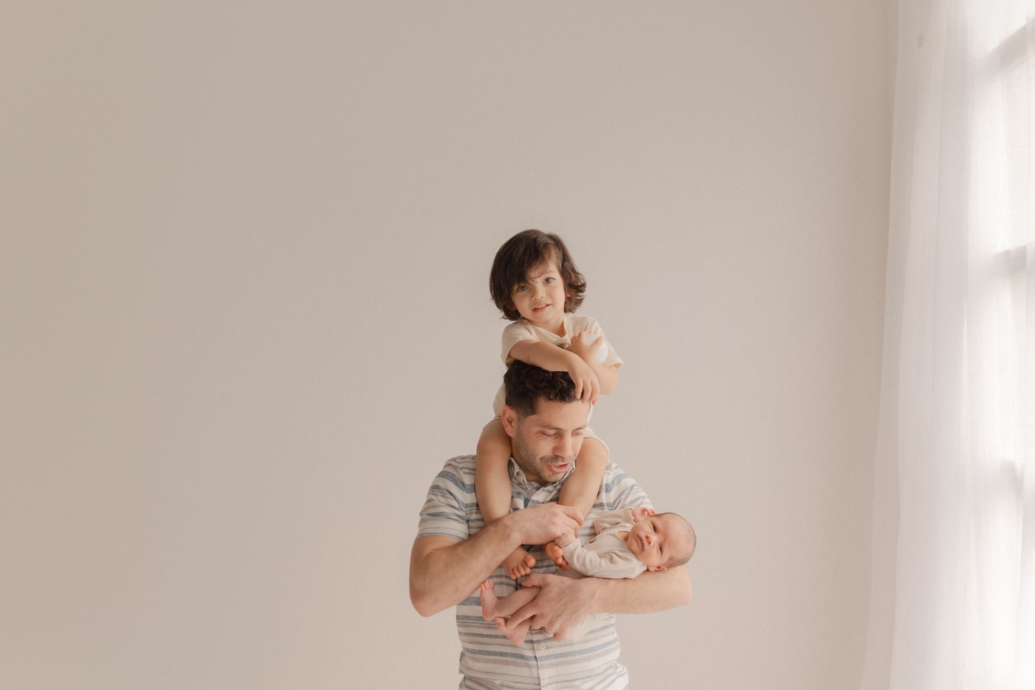 A parent tenderly holds a baby while another child playfully perches on their shoulders in a bright, minimalist setting.