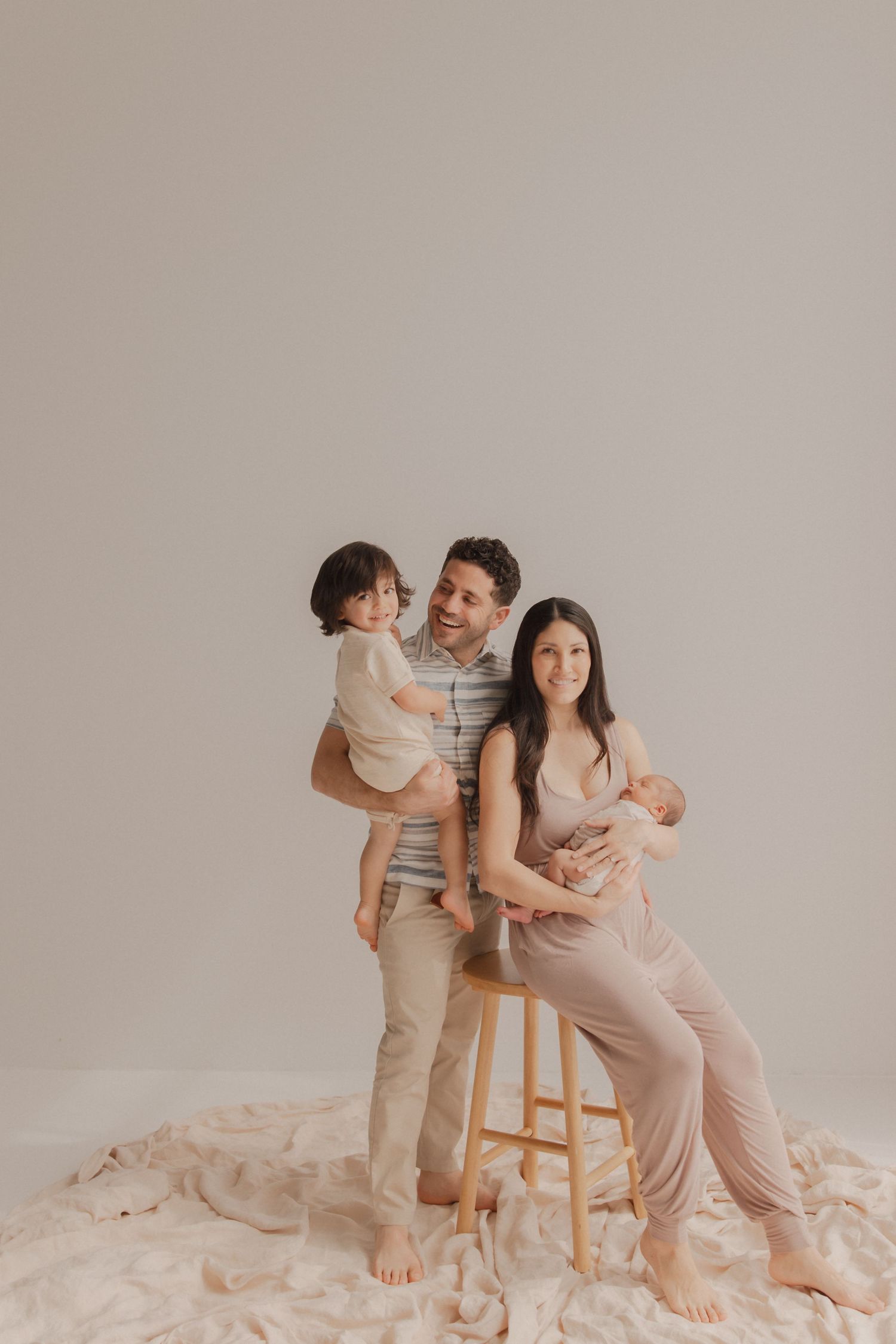 Family poses in neutral tones against beige backdrop, sitting and standing barefoot on soft fabric.