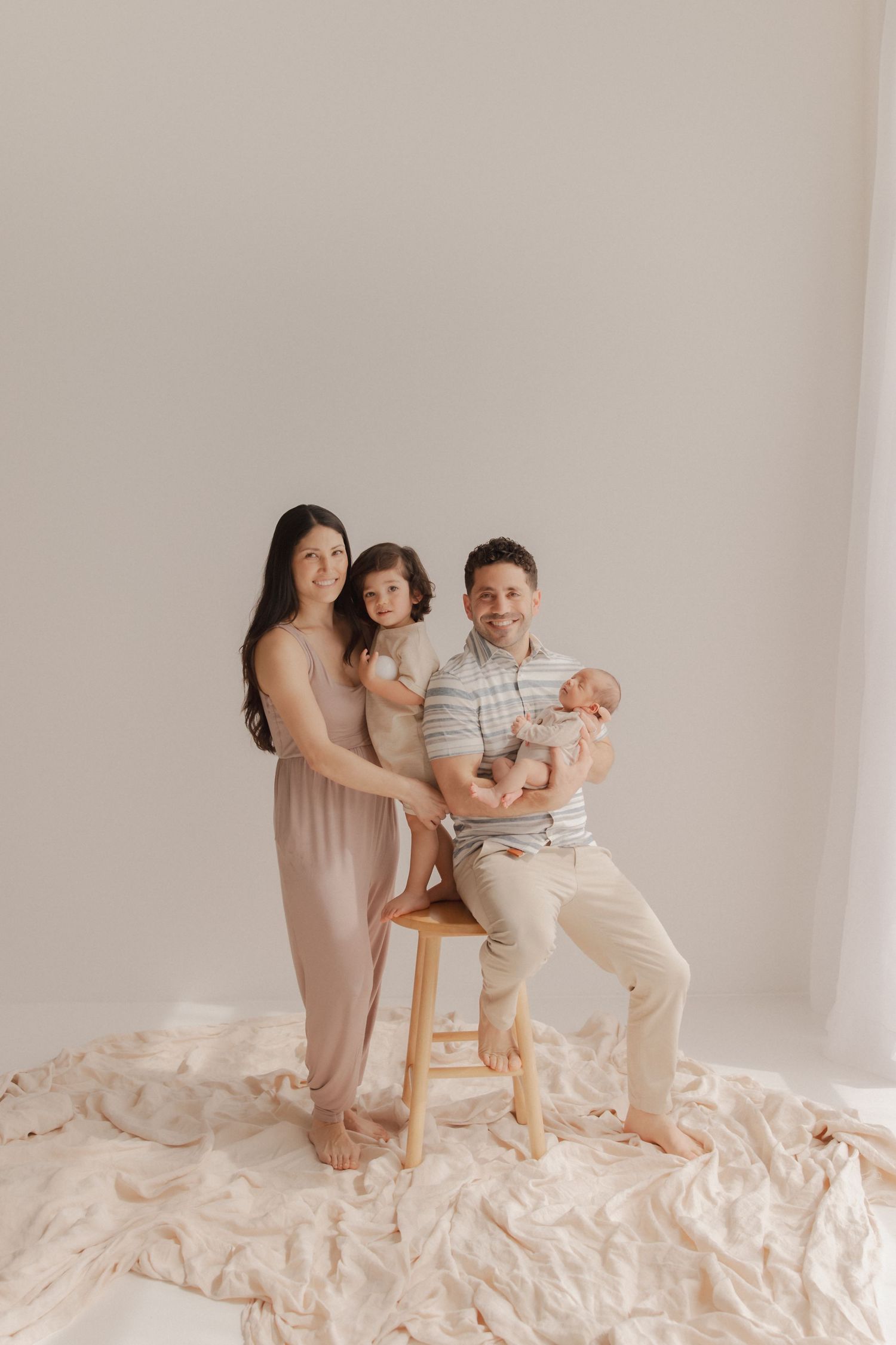 A family poses together on a wooden stool against a neutral backdrop with cream-colored fabric on the floor.