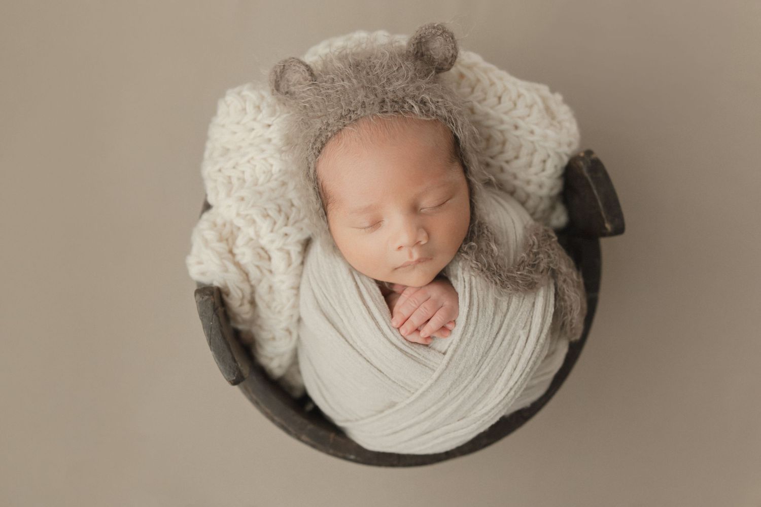 Sleeping newborn baby swaddled in white fabric lying in a dark bowl with knitted accessories and bear ear hat.