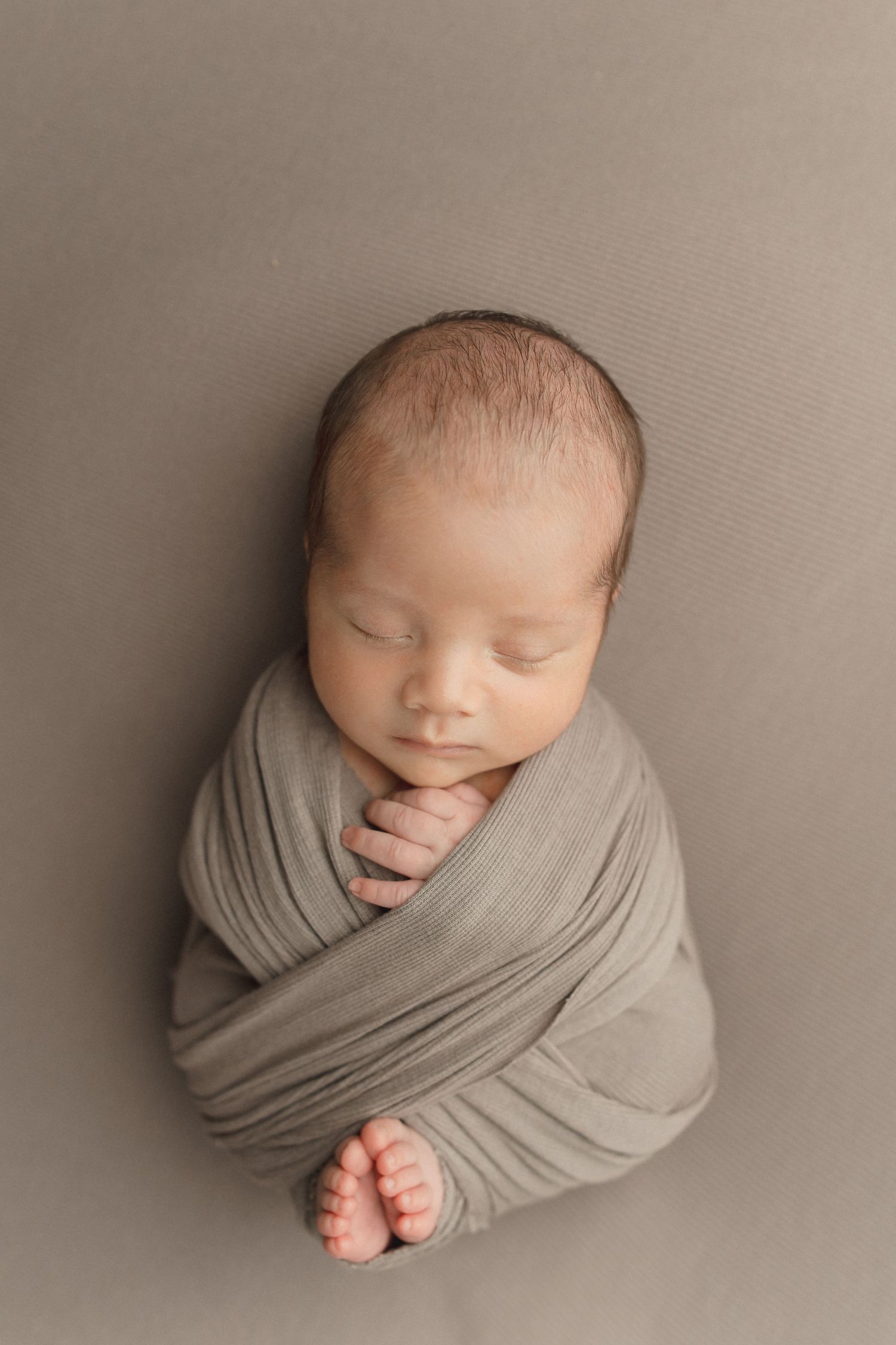 Newborn baby swaddled in soft gray fabric wrap on neutral backdrop with tiny feet peeking out.