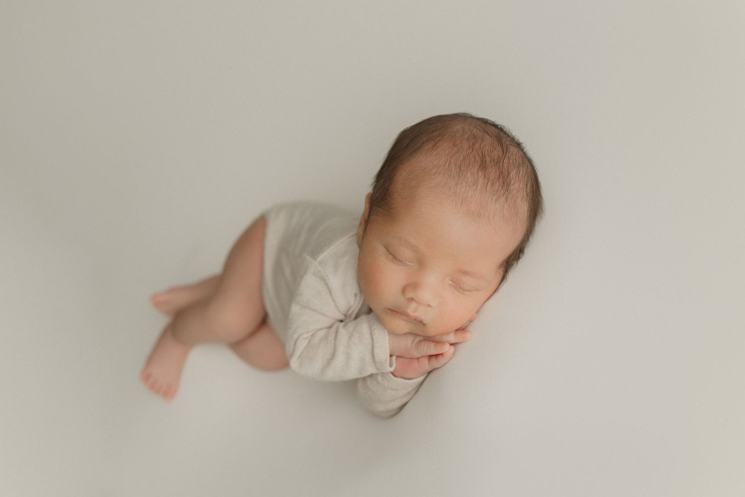 Peaceful sleeping newborn baby wearing cream outfit lying on soft beige backdrop during professional photoshoot.