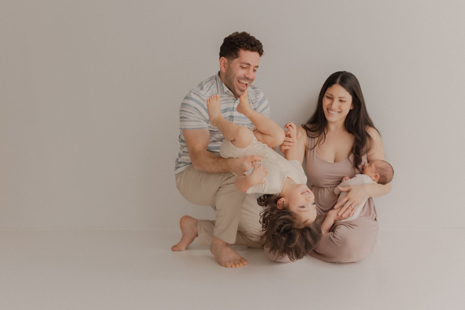 A family laughs and plays together during a minimalist studio photo session against a neutral backdrop.