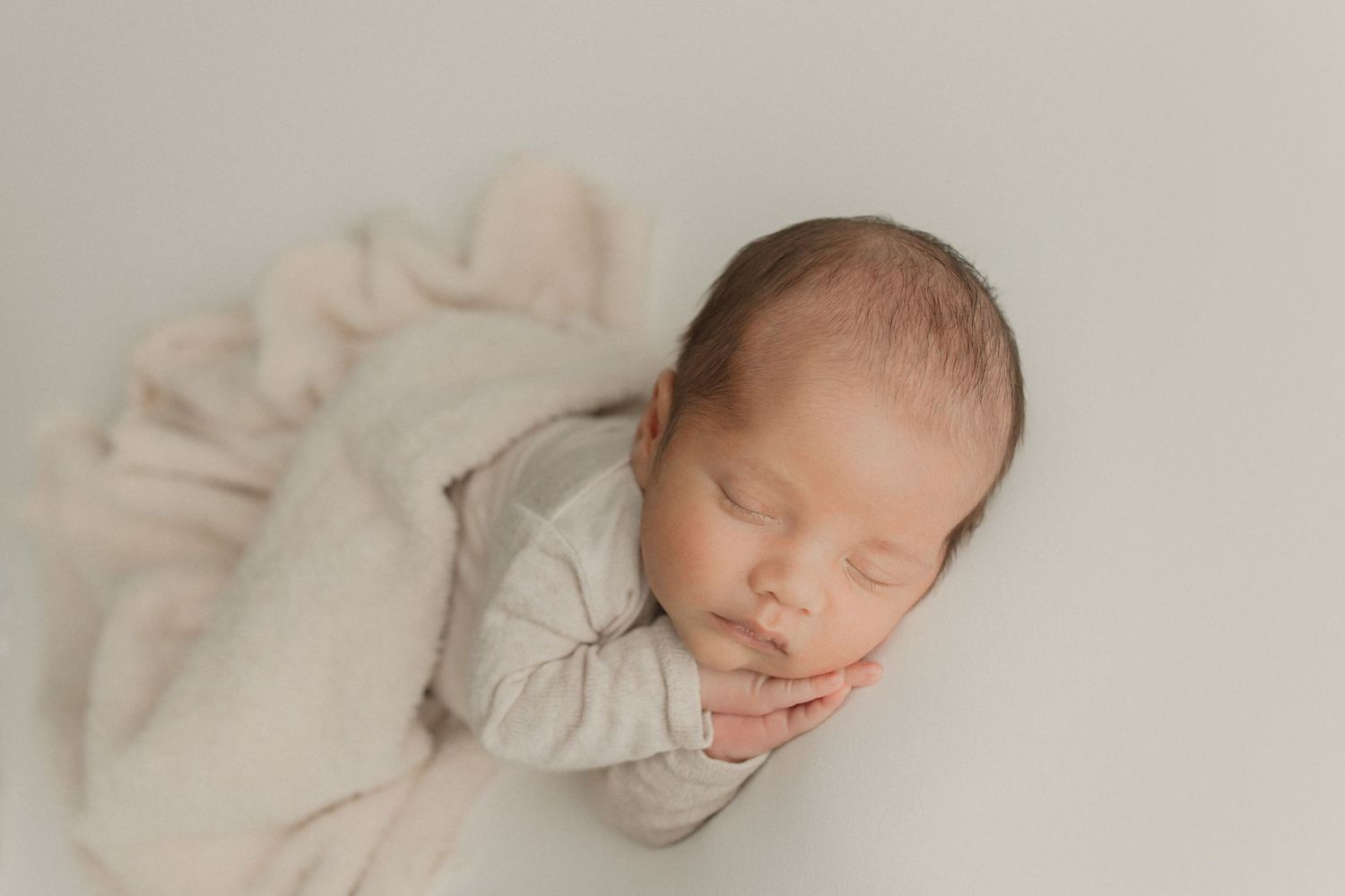 Sleeping newborn wearing light colored outfit lying on soft cream fabric background.