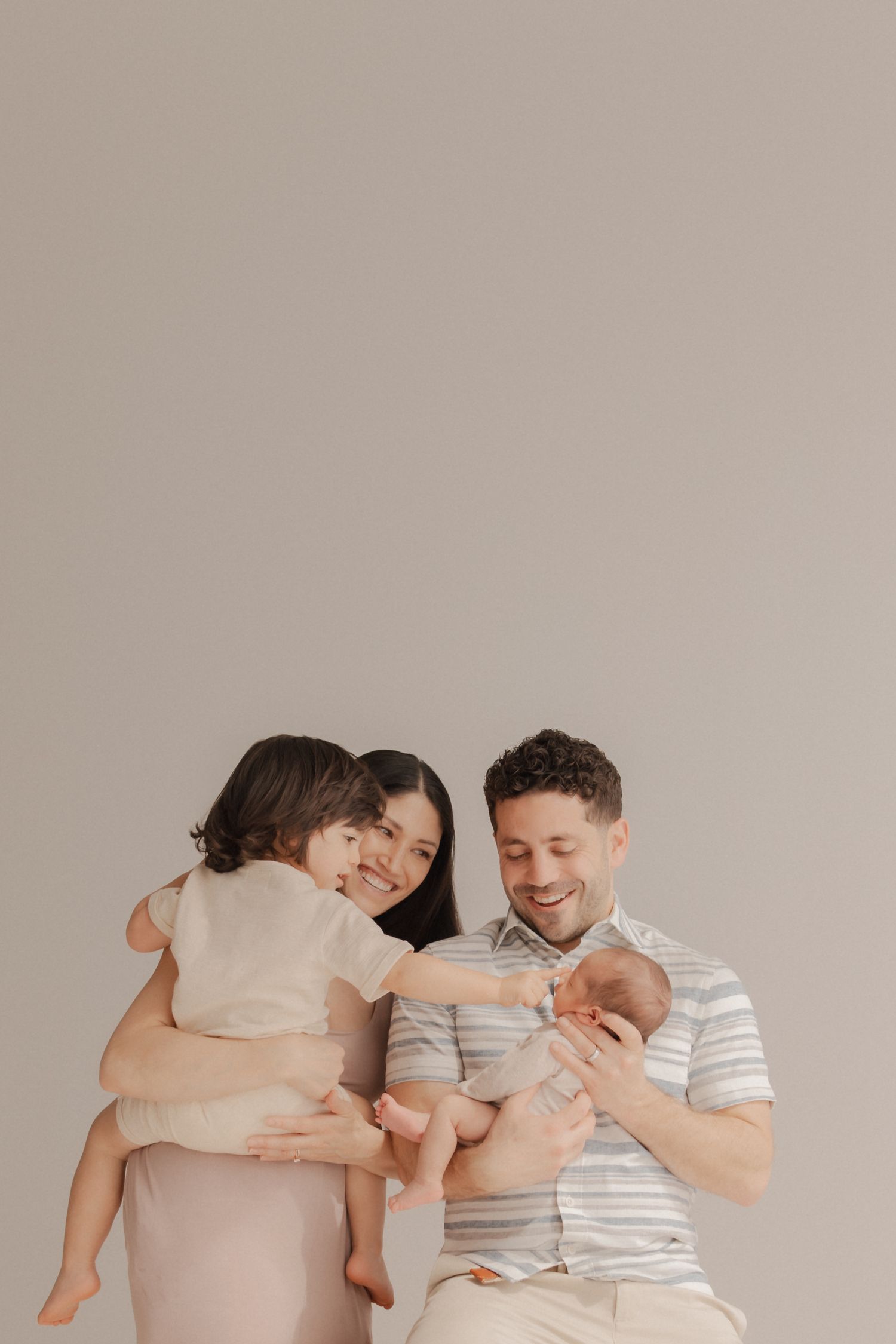 A serene family photograph in muted neutral tones showing parents holding two small children against a beige background.