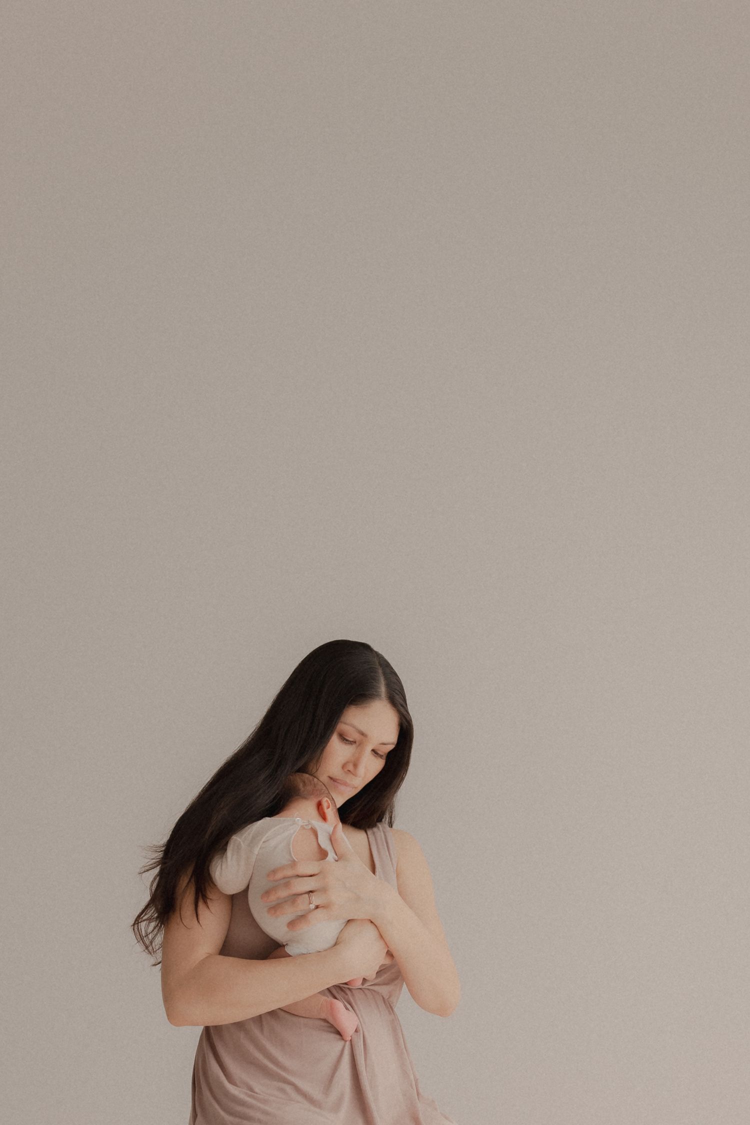 Mother in pink dress tenderly embraces and nurtures her baby against neutral beige background.