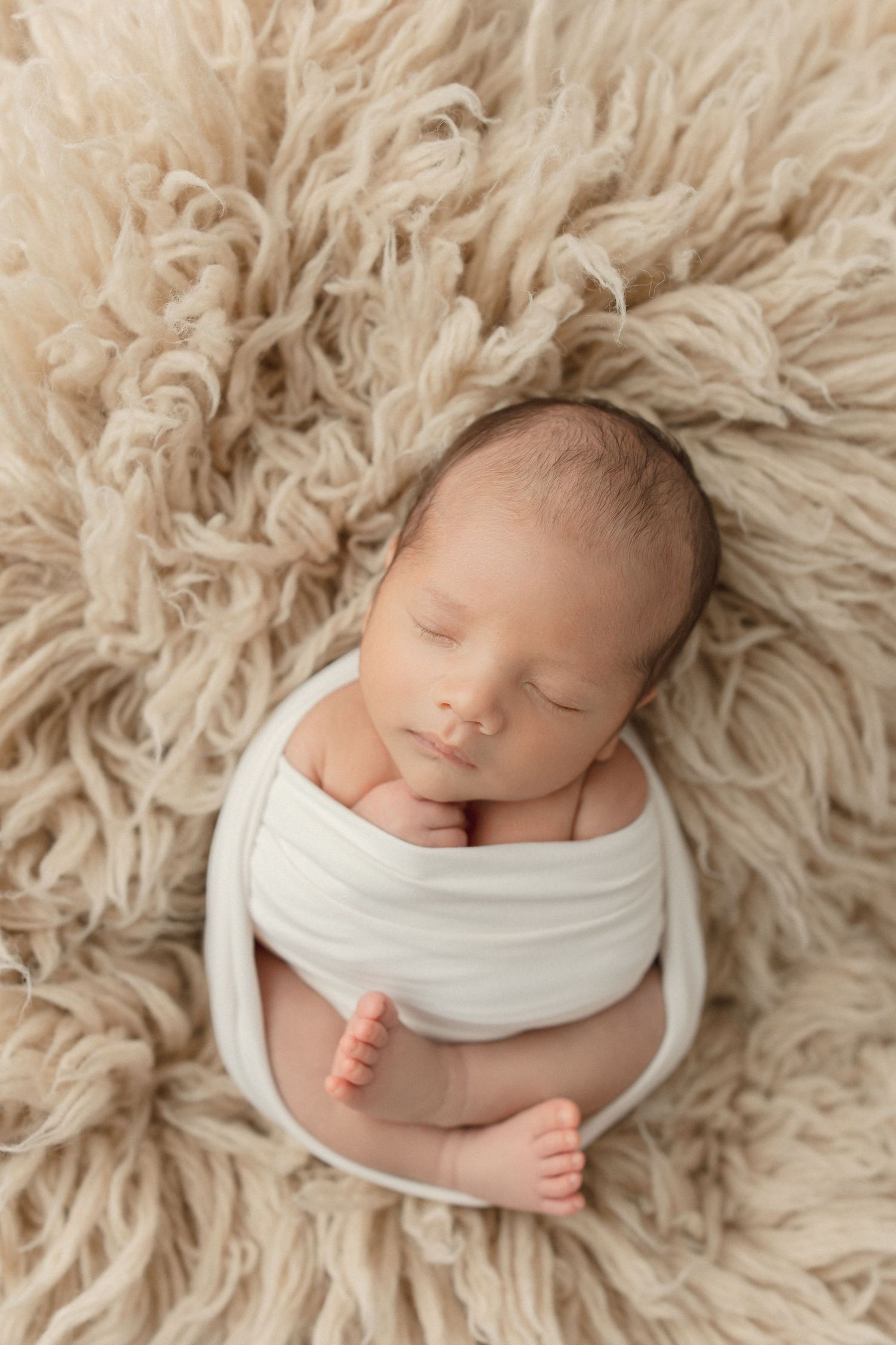 Sleeping newborn wrapped in white fabric lying on soft beige furry blanket during professional photo session.