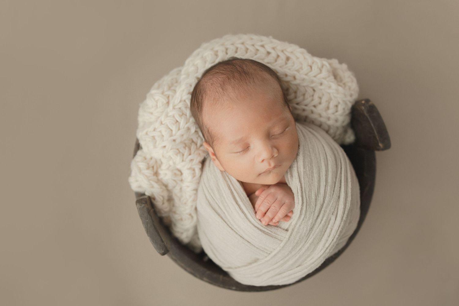 Sleeping newborn baby wrapped in white fabric and wearing knitted hat rests in round bowl during professional photo session.