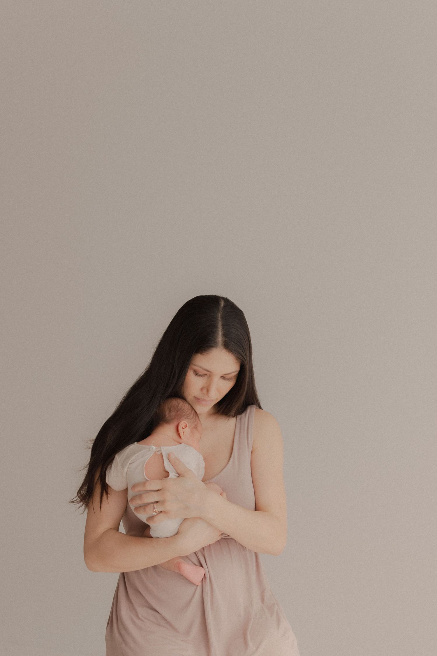 A woman in beige dress holds and looks down tenderly at an infant against neutral backdrop.
