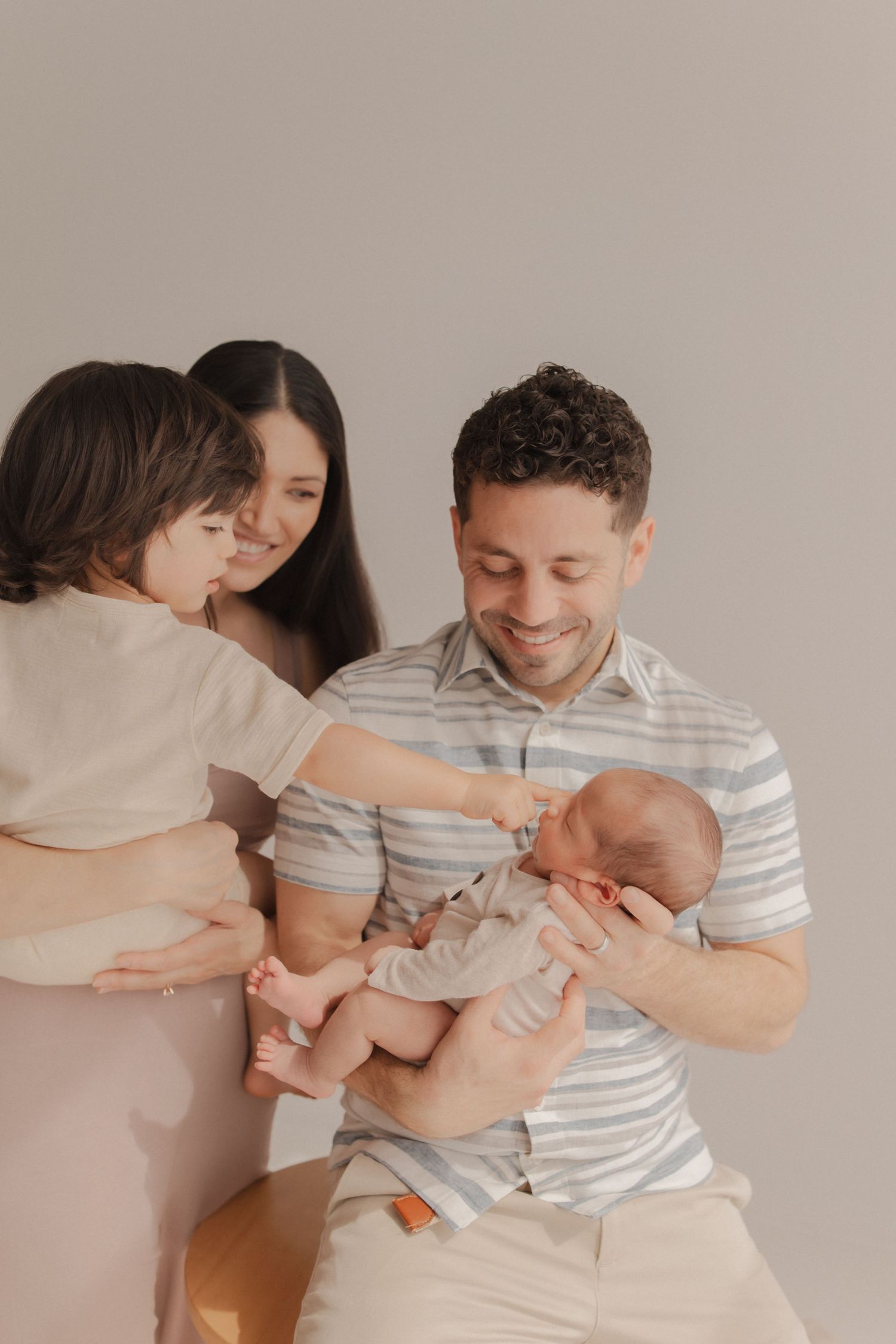 Family shares tender moment with newborn baby in cozy indoor setting wearing neutral colored clothing.