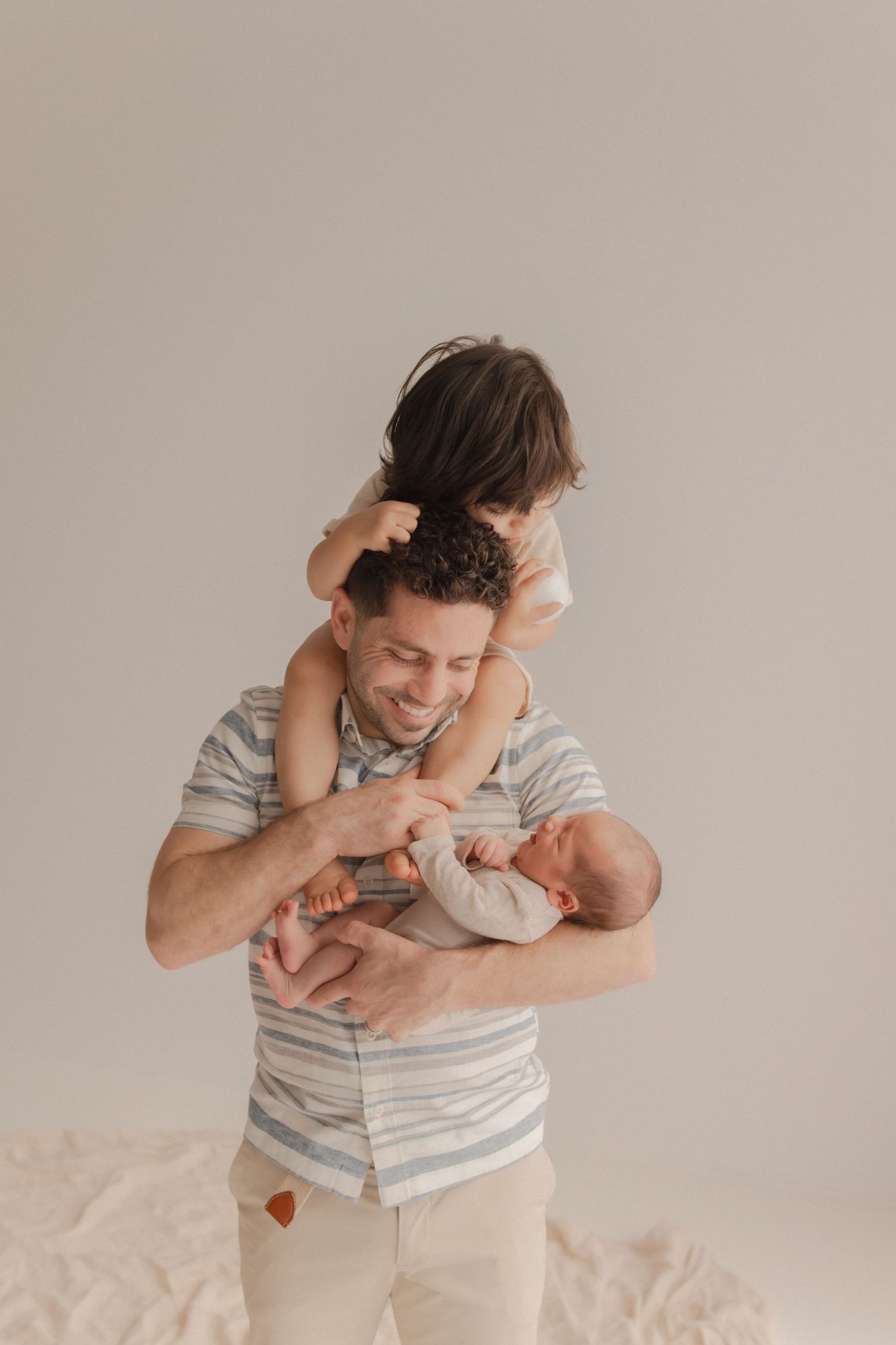 A parent carries a small child on their shoulders while both laugh and play together against a neutral backdrop.