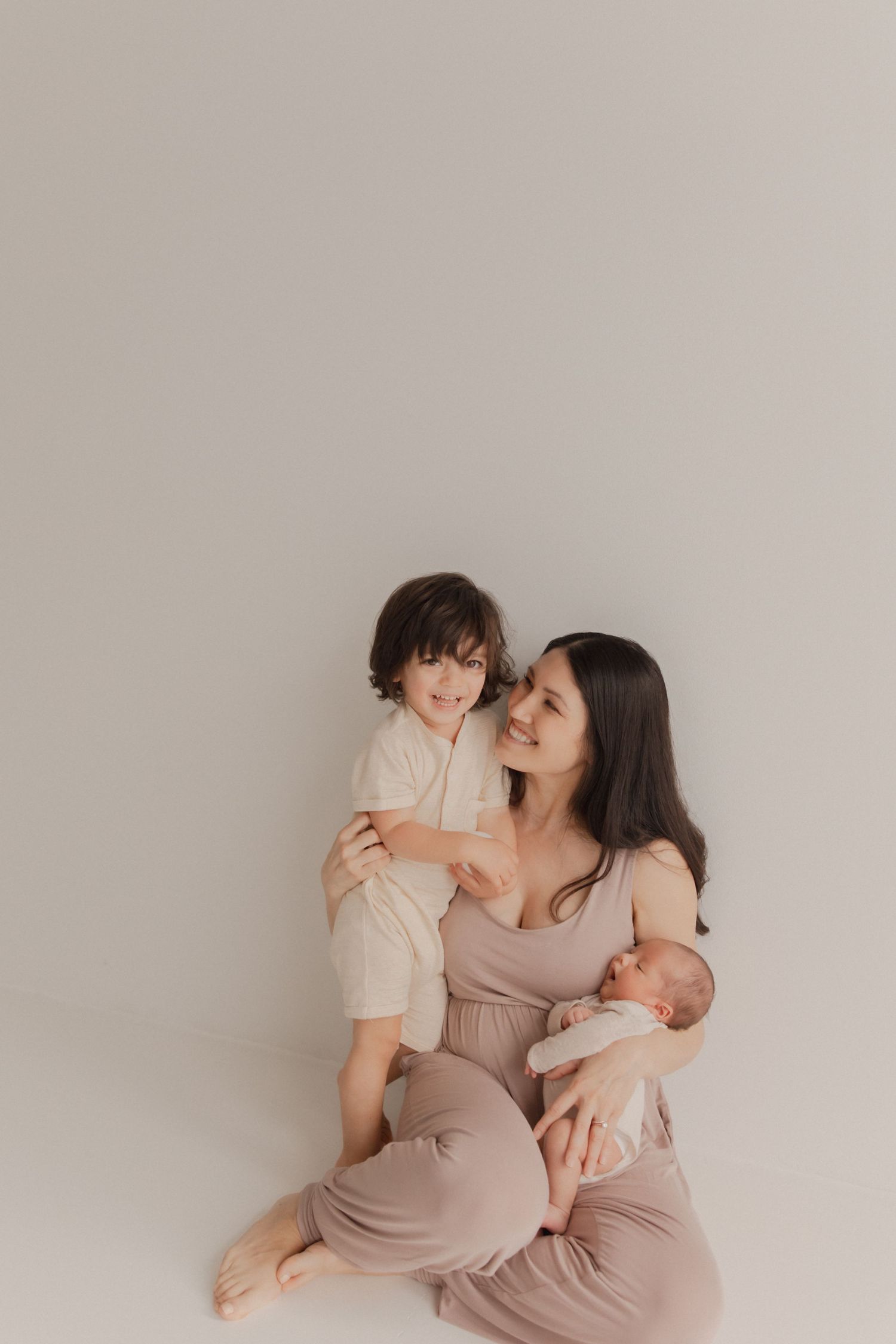Loving family portrait of mother in beige dress holding baby while embracing toddler against neutral background.