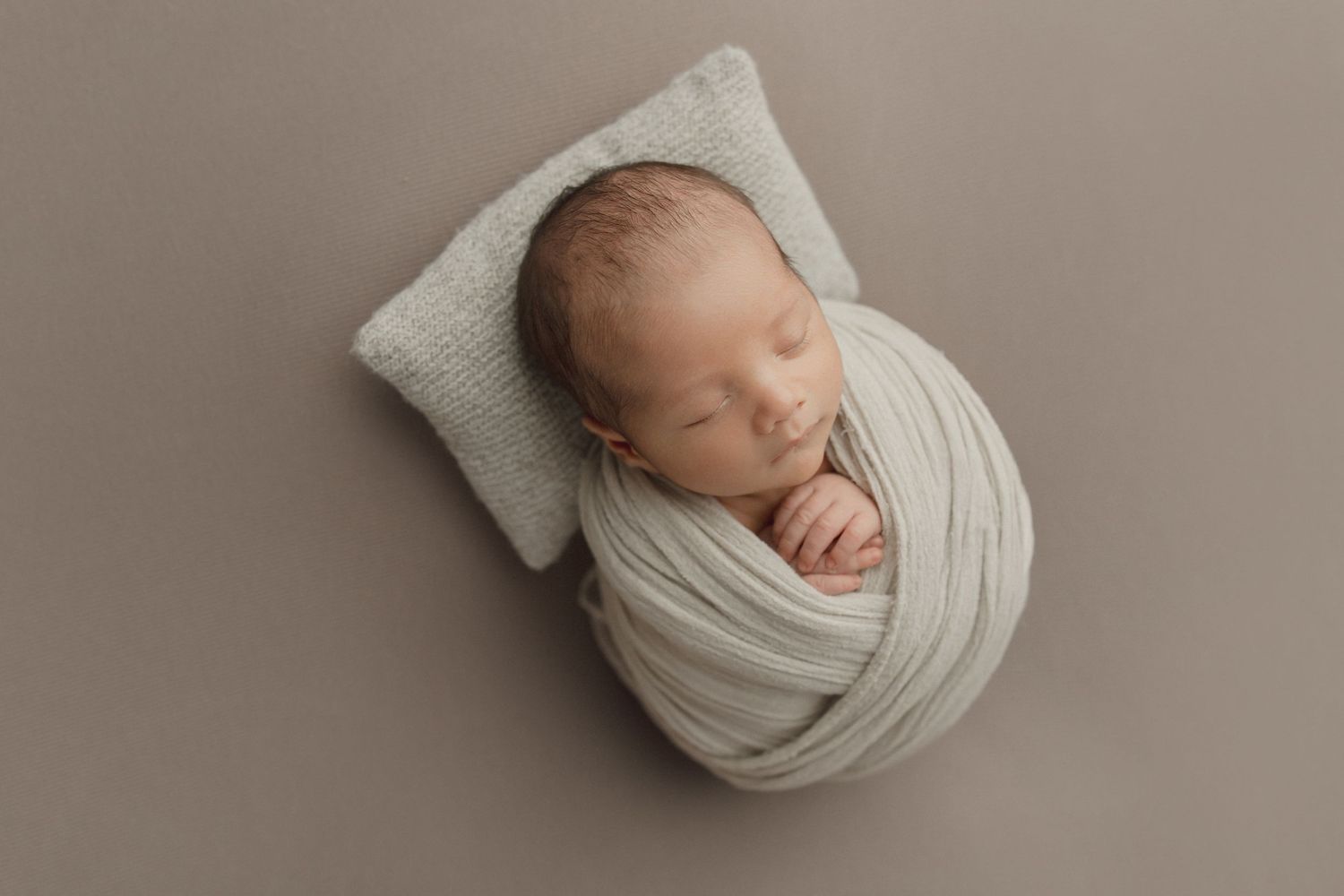 A sleeping newborn baby wrapped snugly in cream-colored fabric rests peacefully on a soft pillow against a neutral backdrop.