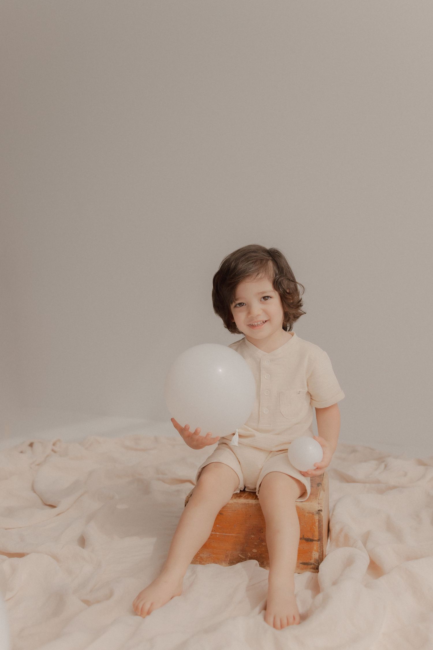 Young child in cream colored outfit sitting on blanket holding white balloon against neutral background.