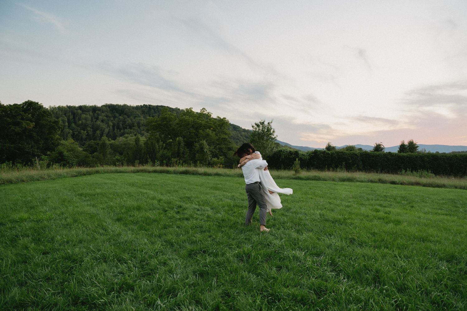 A couple embracing and dancing in an open field with mountains in the distance at sunset.