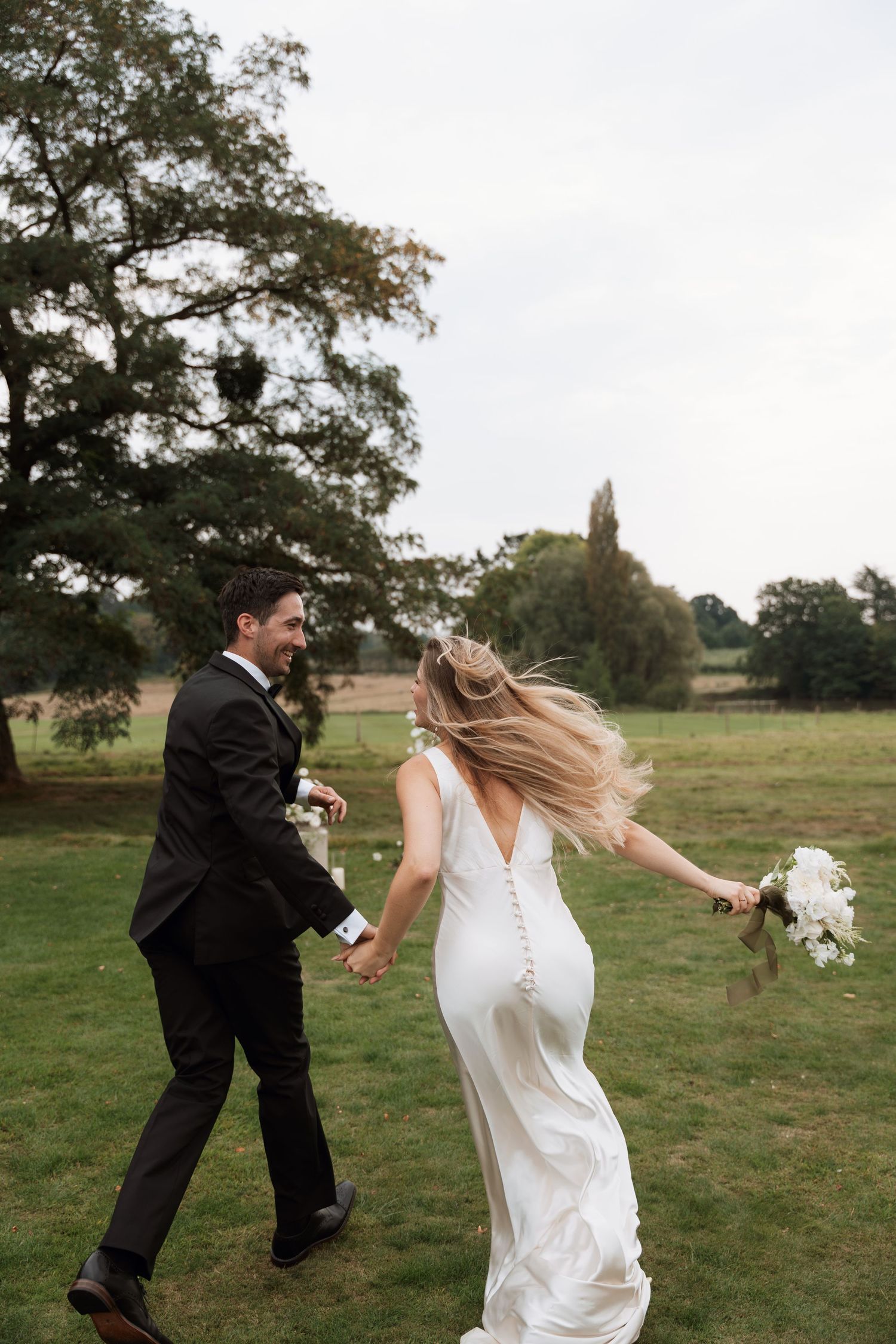 A romantic moment between newlyweds dancing playfully on a grassy field with a church steeple visible in the background.