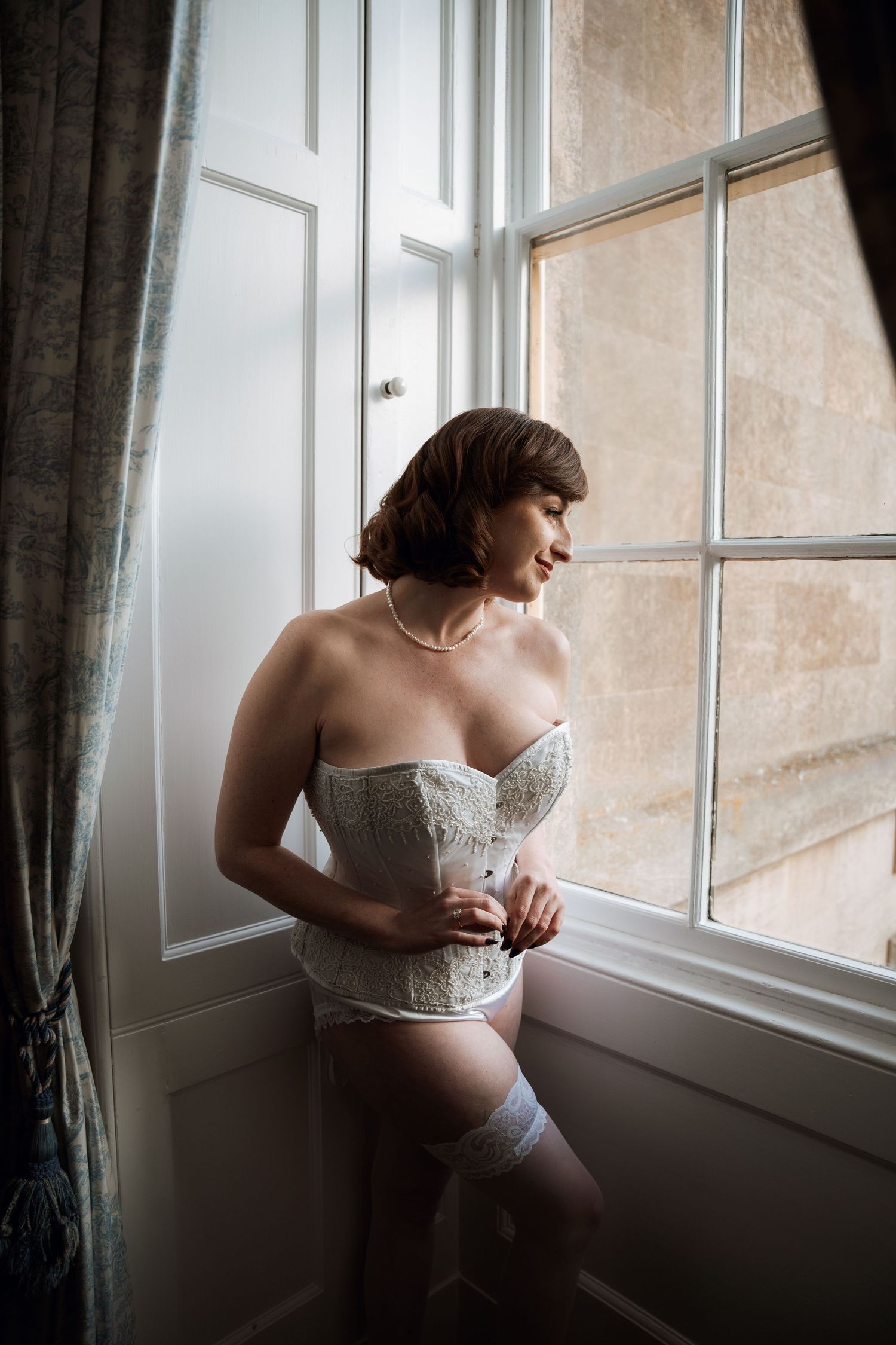 Woman in white lace lingerie posing by window in intimate boudoir photo.
