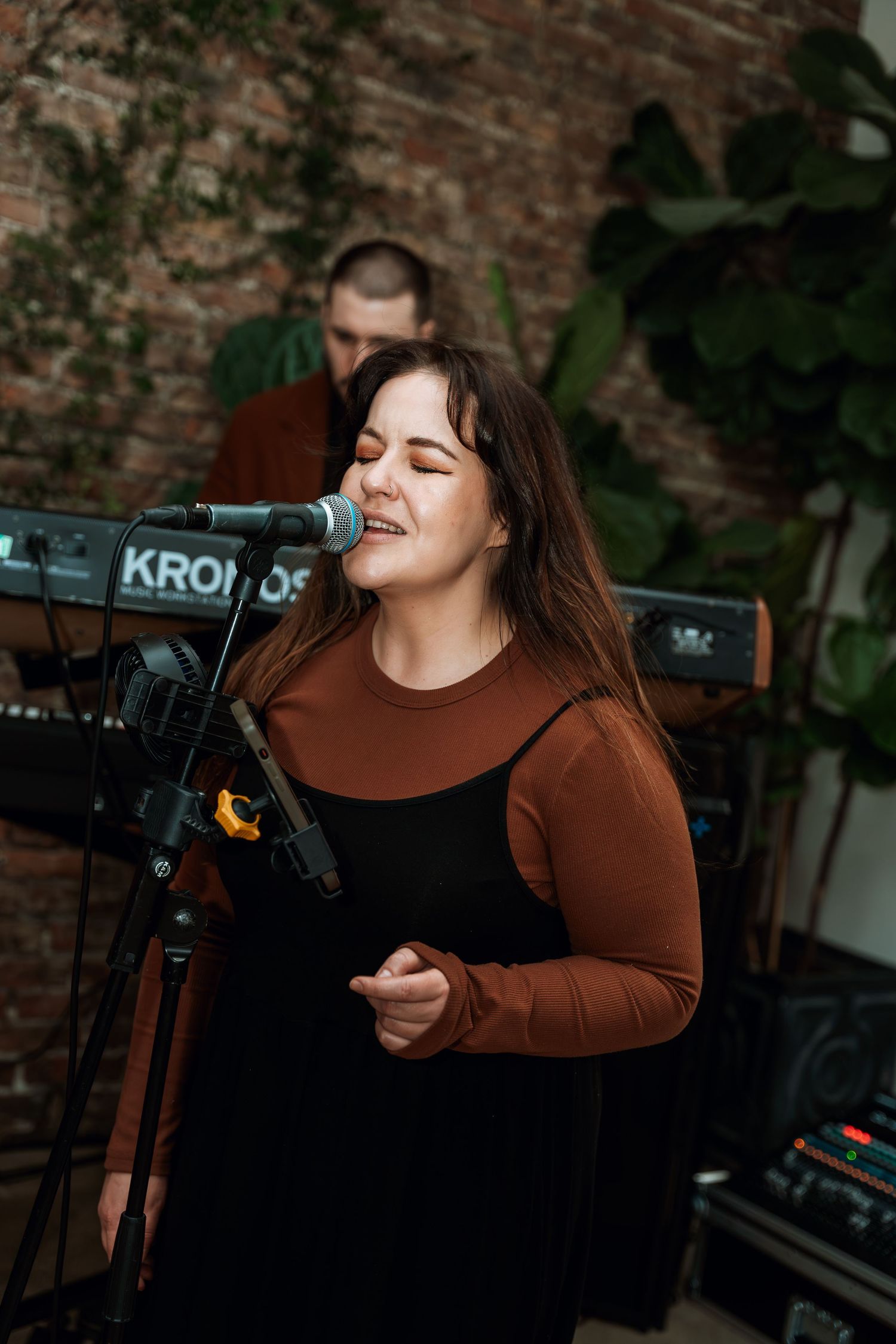 Performer singing into microphone with Korg keyboard against brick wall background.