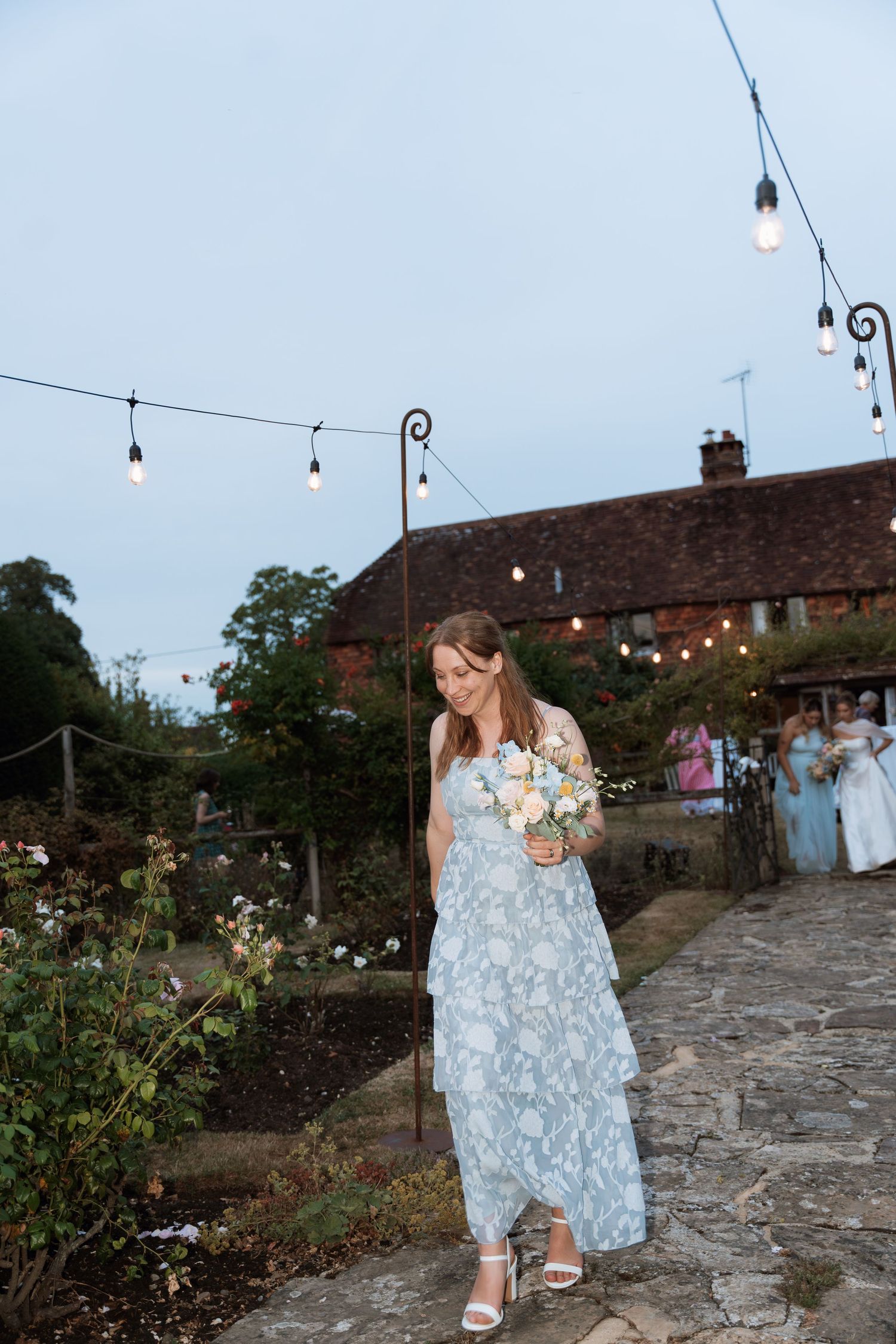 A guest walking through a garden path with string lights and historic brick building in background.