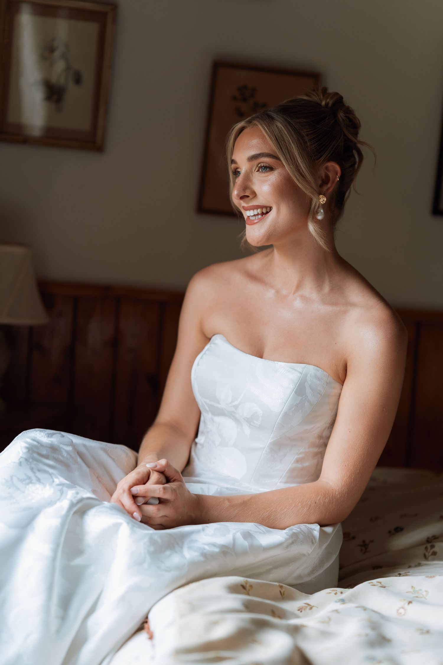 A bride in a white strapless wedding dress sits on a bed by a window smiling with her hair elegantly styled.