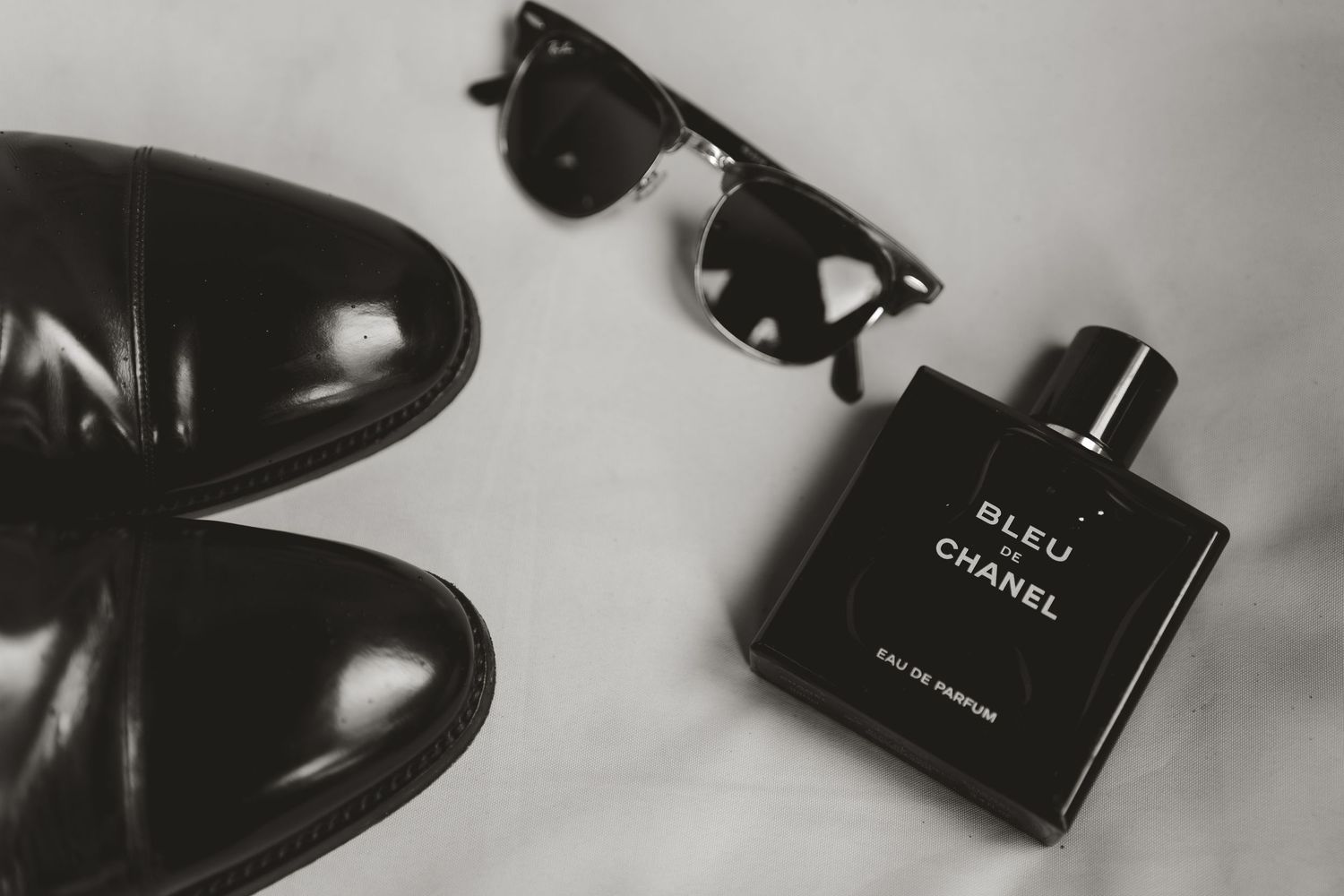 Monochromatic still life with black leather shoes, sunglasses, and Chanel Bleu cologne bottle on light background.