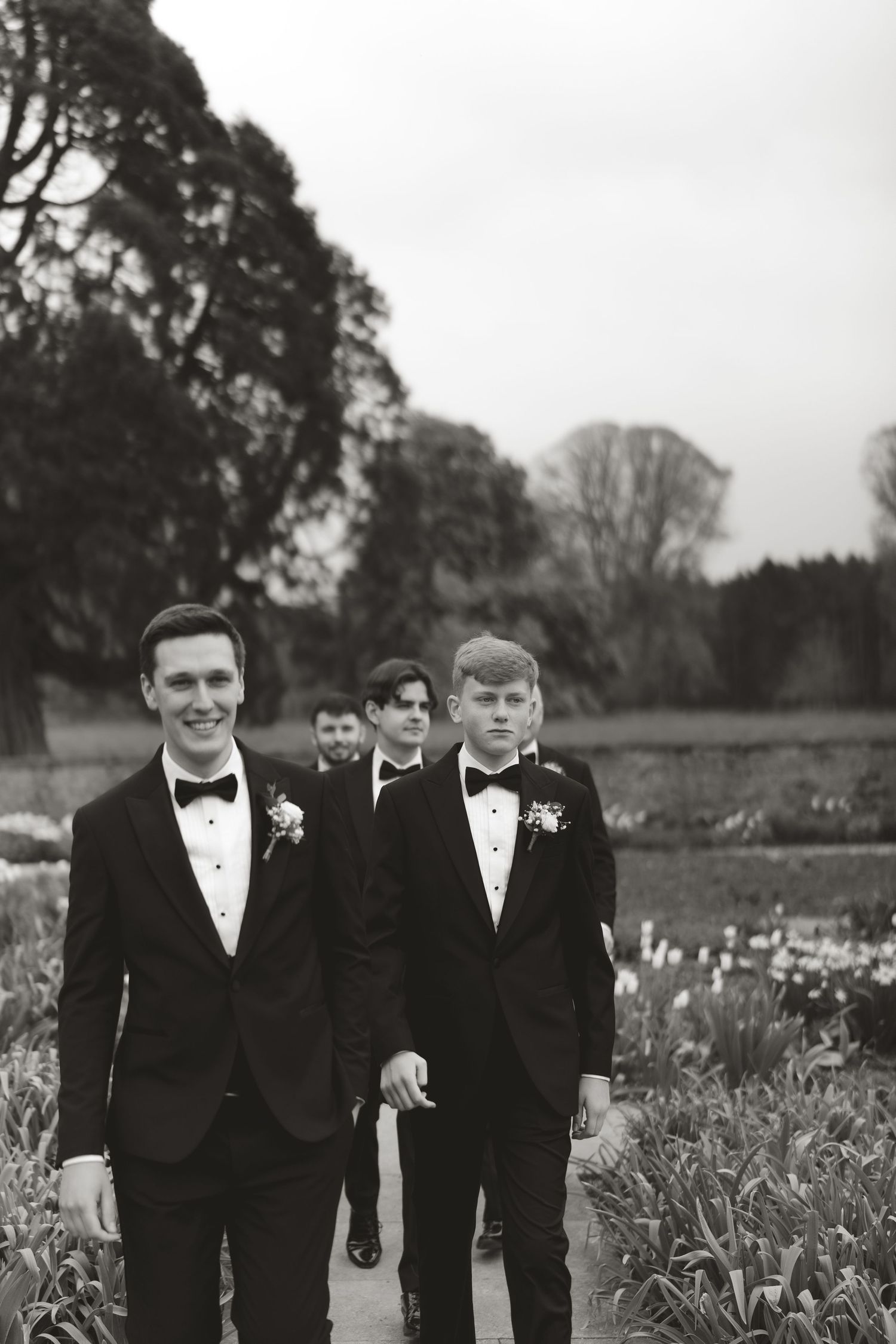 Groomsmen in black tuxedos and bow ties walking together through a garden setting.