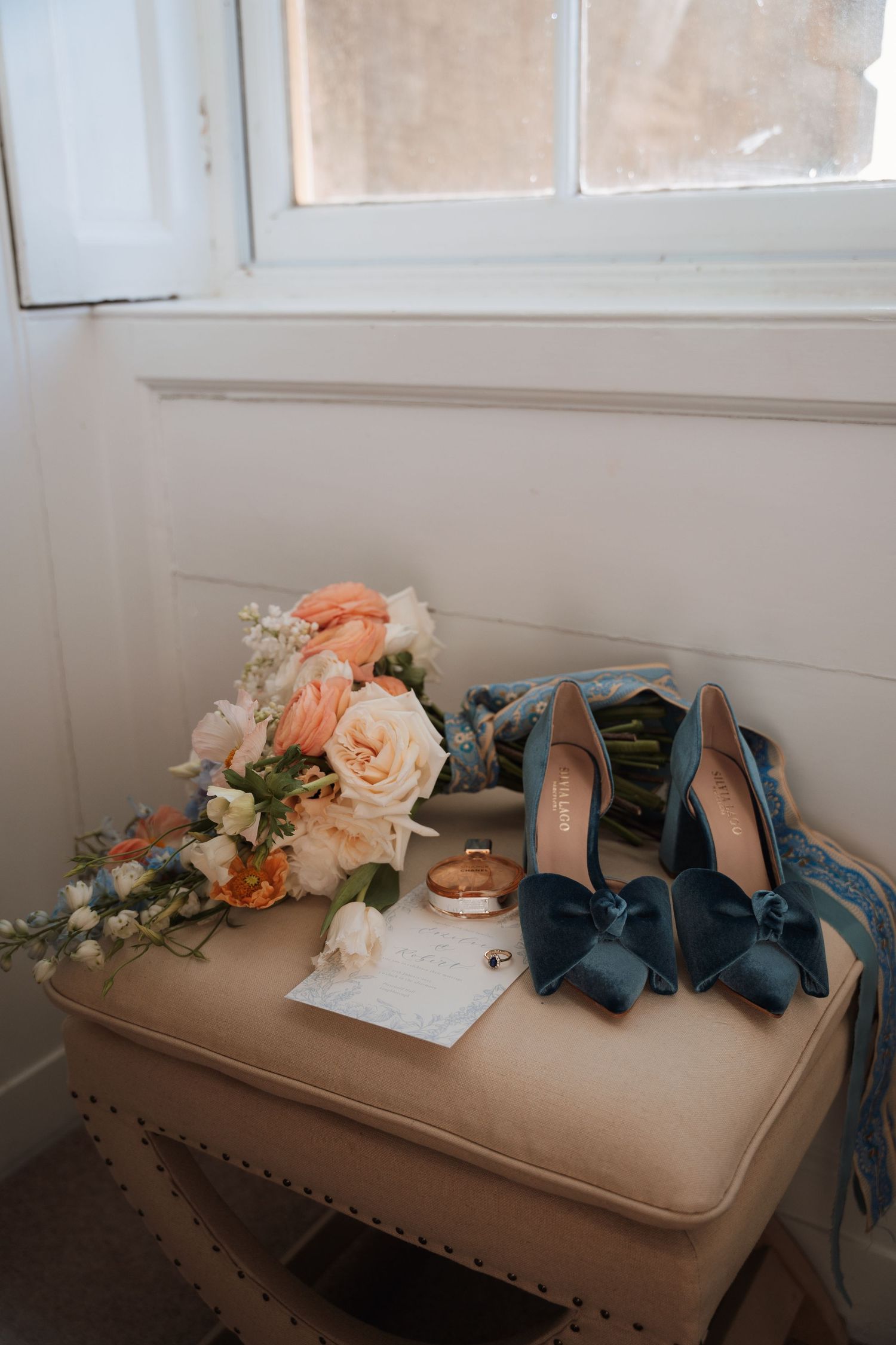 Wedding details including blue velvet shoes, rings, and peach roses arranged on a beige ottoman.
